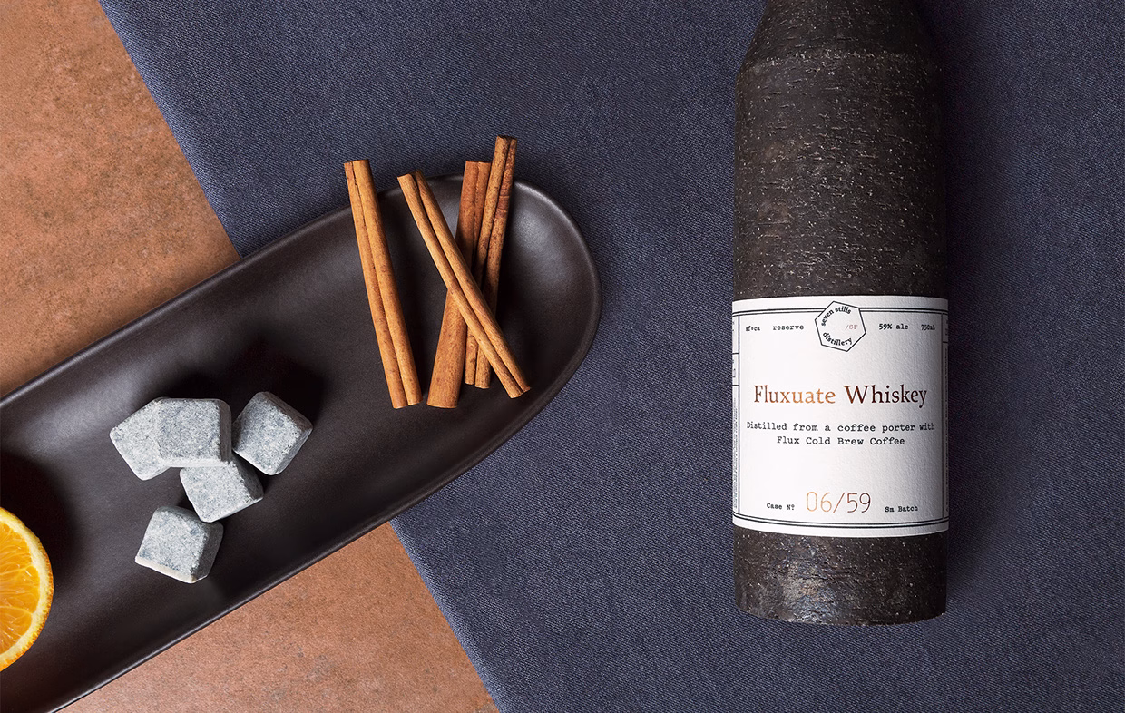 A dark bottle of Fluxuate Whiskey rests next to a tray of cinnamon sticks and gray whiskey stones, on textured fabrics.