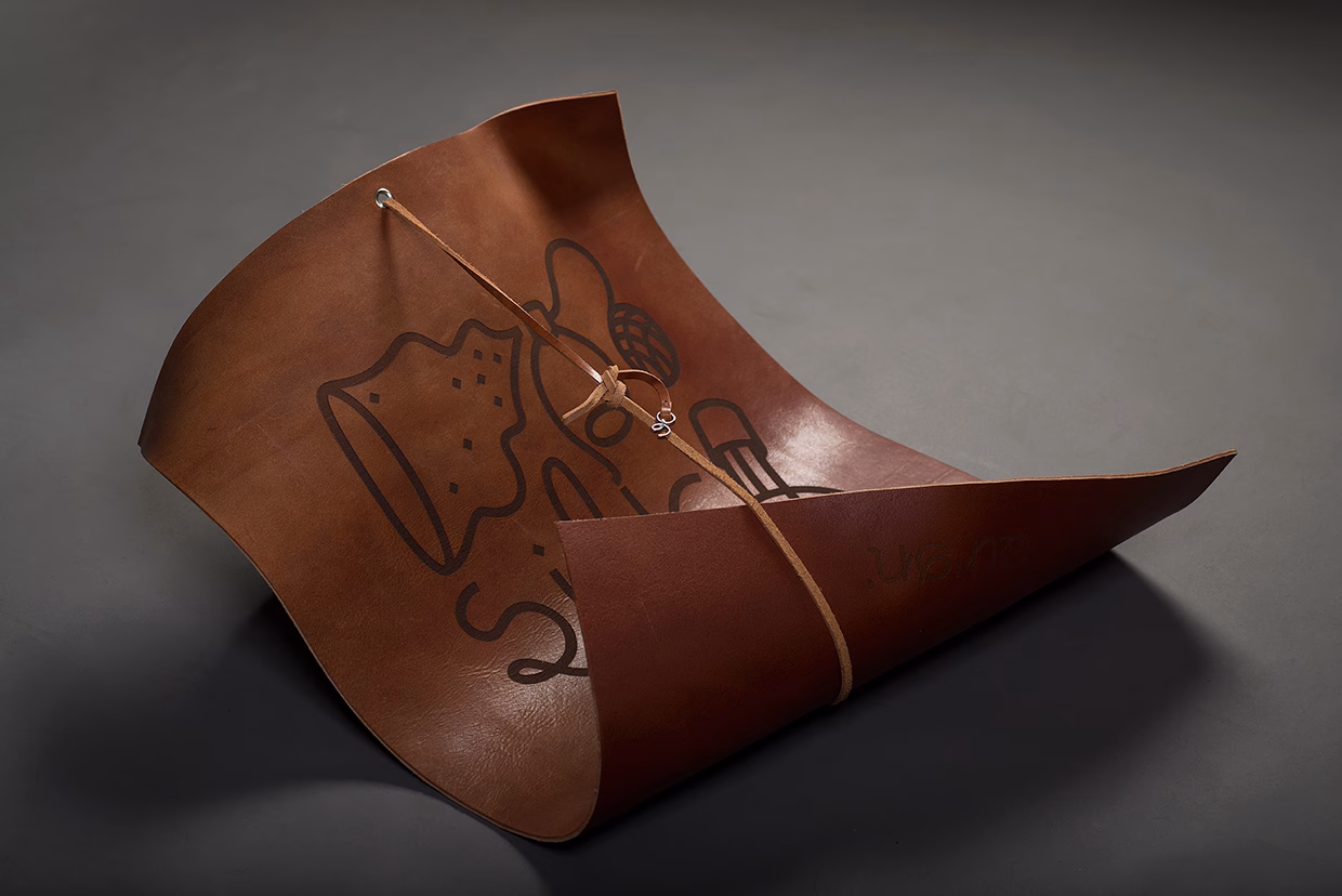 A curved piece of brown leather with a graphic design, secured with a thin strap, resting on a dark surface.