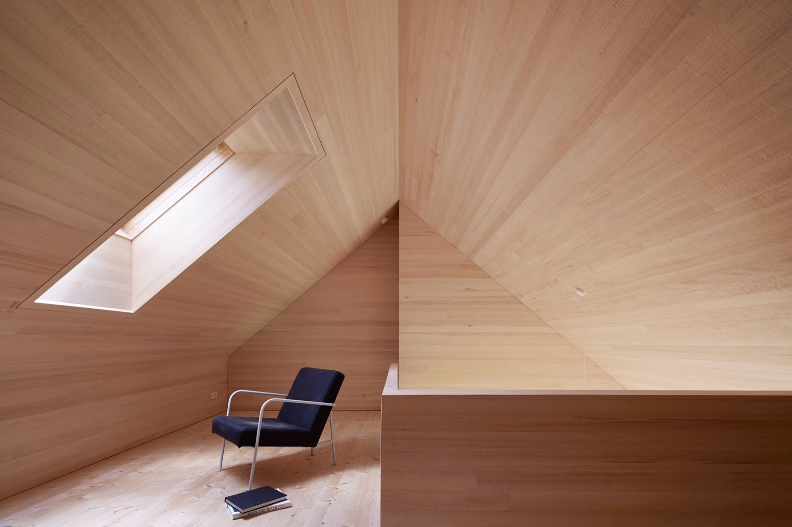 A minimalist room features pale wooden walls and a slanted ceiling, with a black chair and skylight casting light inside.