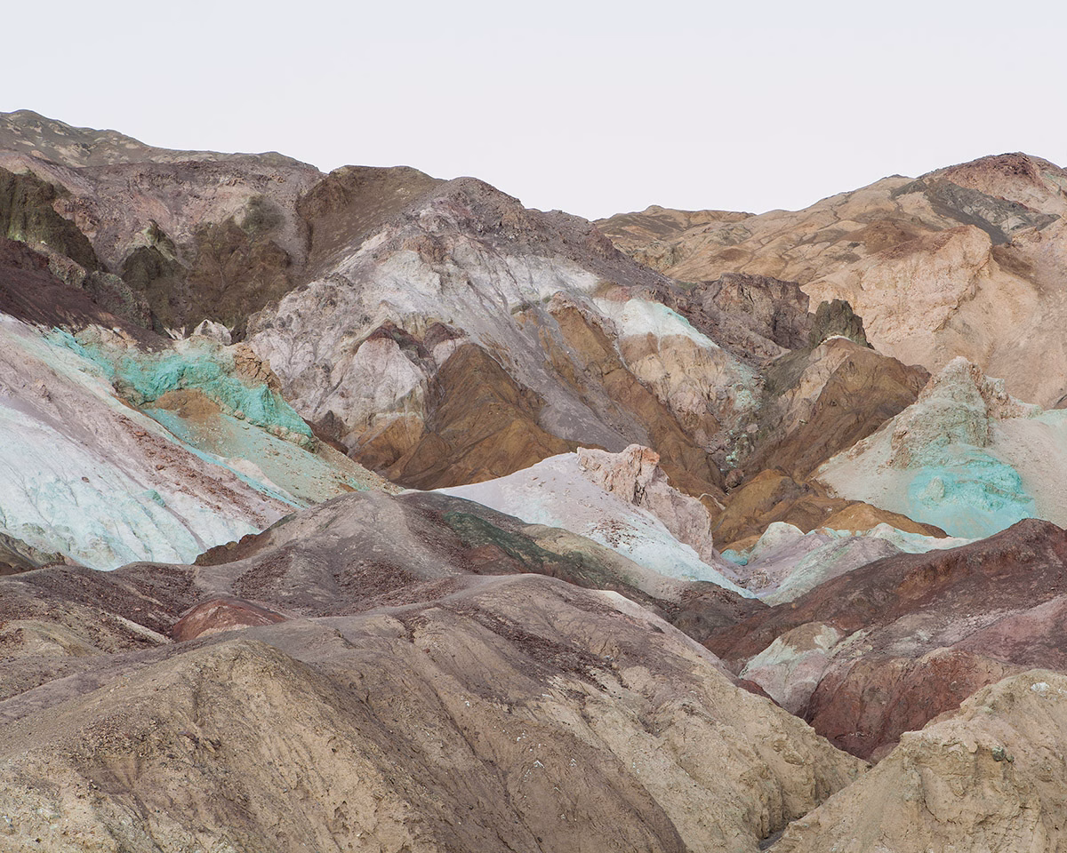 Colorful, layered mountain ridges display earthy tones of ochre, green, and gray under a clear sky.