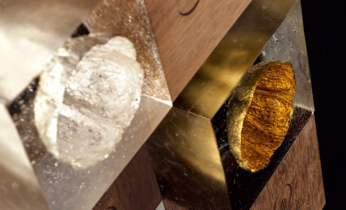 Close-up of two hexagonal forms, one in clear resin and one in shiny gold, mounted on a wooden base.