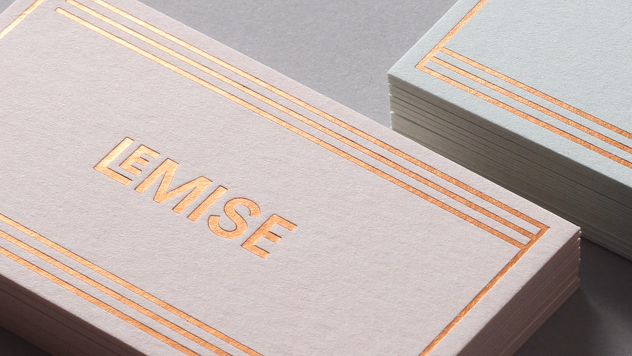 A close-up of two business cards features muted colors and embossed copper detailing, one pale gray and one light mint.