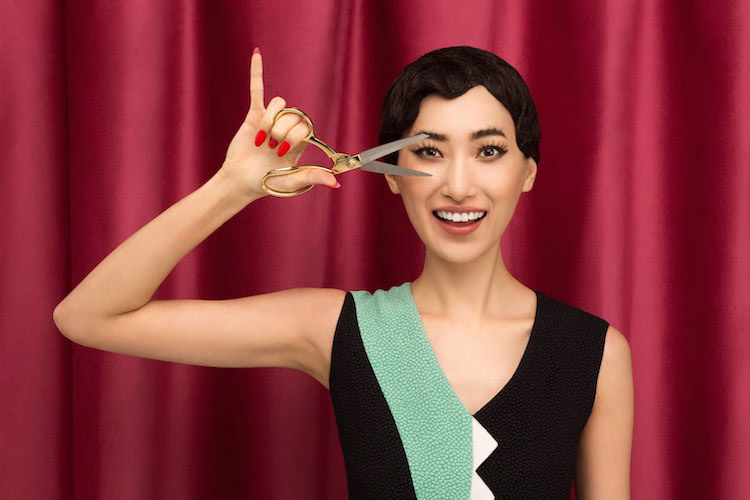 A woman with short hair smiles while holding a pair of gold scissors against a vibrant red curtain.