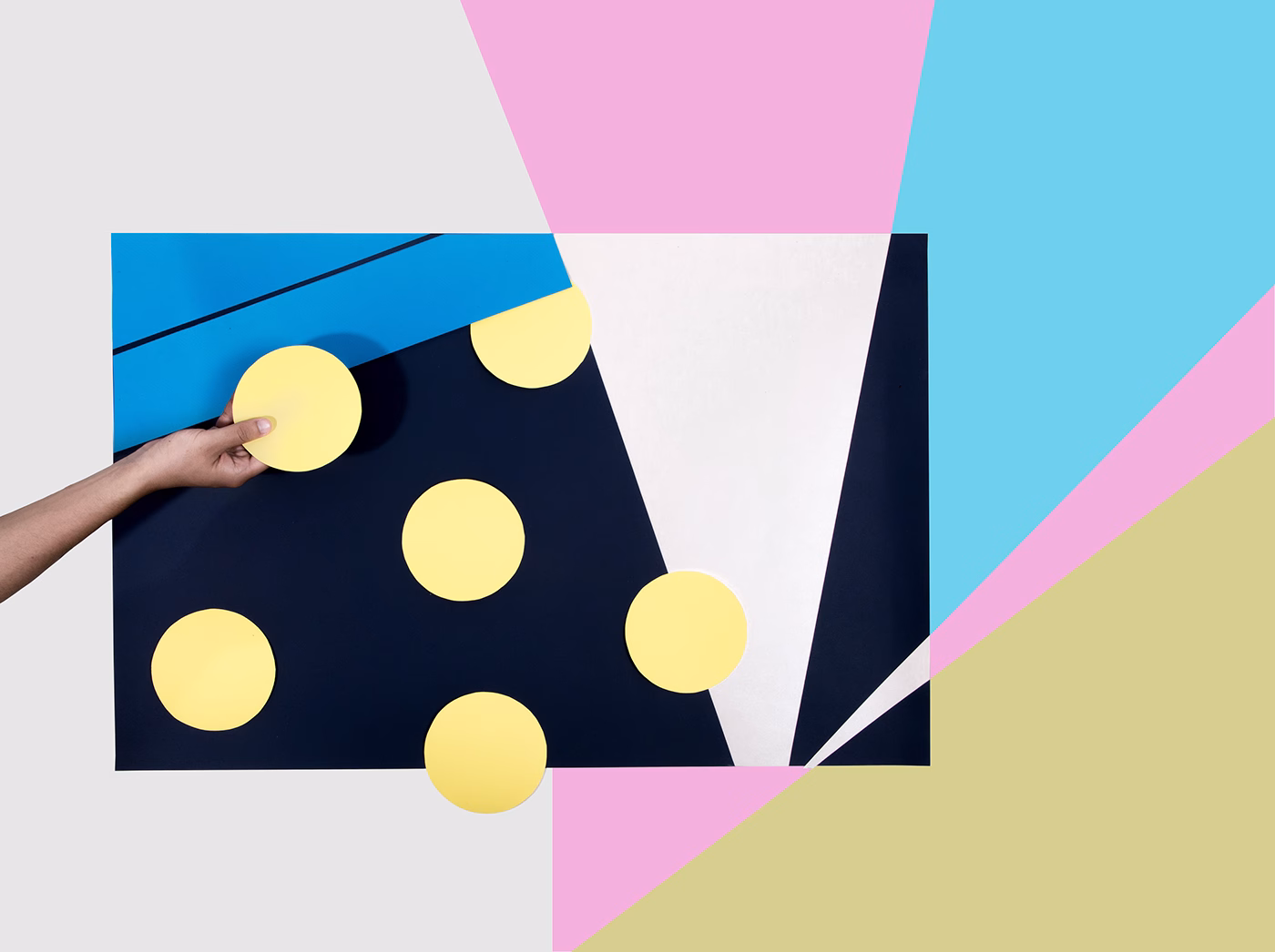 A hand places a yellow circle on a navy background, surrounded by colorful triangular shapes in various pastel tones.