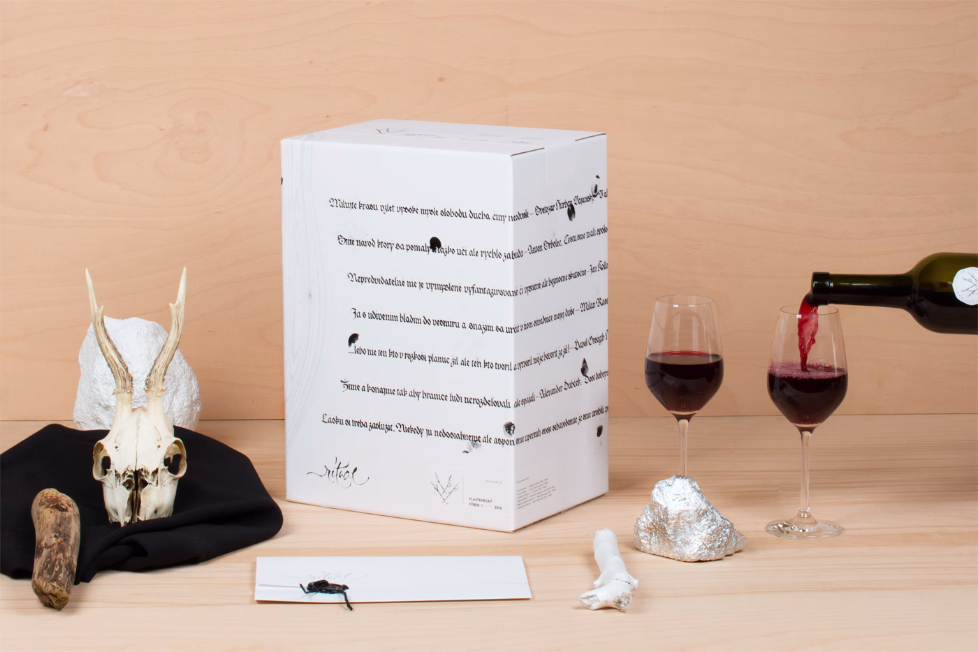 A white wine box with black typography sits near two filled glasses, a skull, and other textured elements on wood.