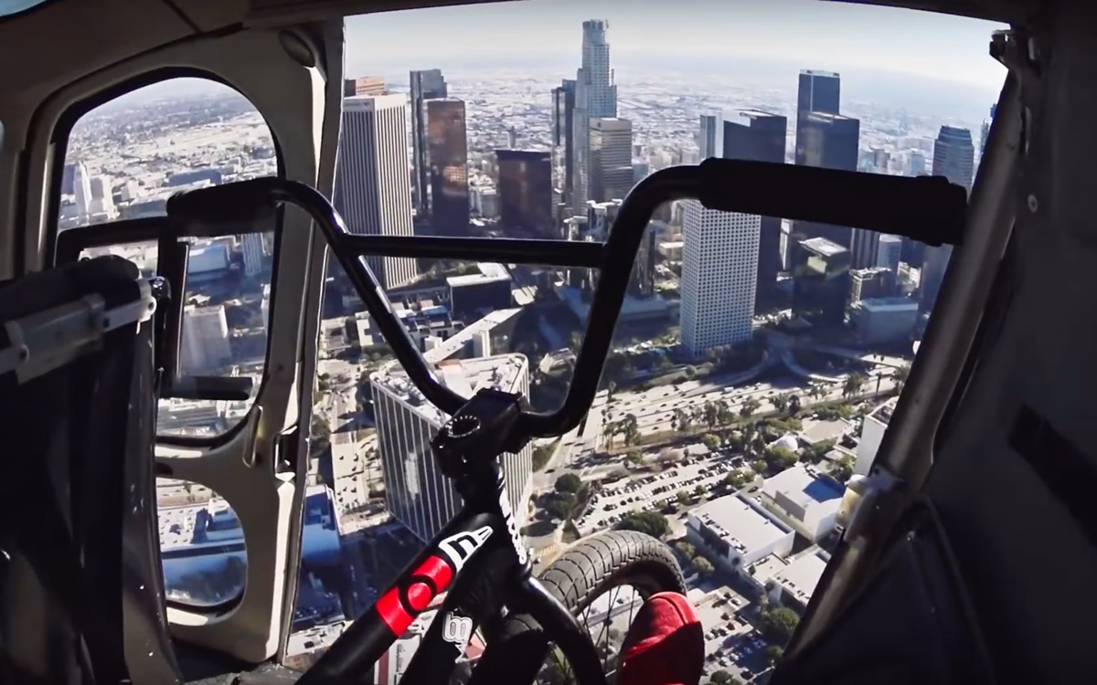 A view from a helicopter shows a BMX bike's handlebars in the foreground, with the Los Angeles skyline below.