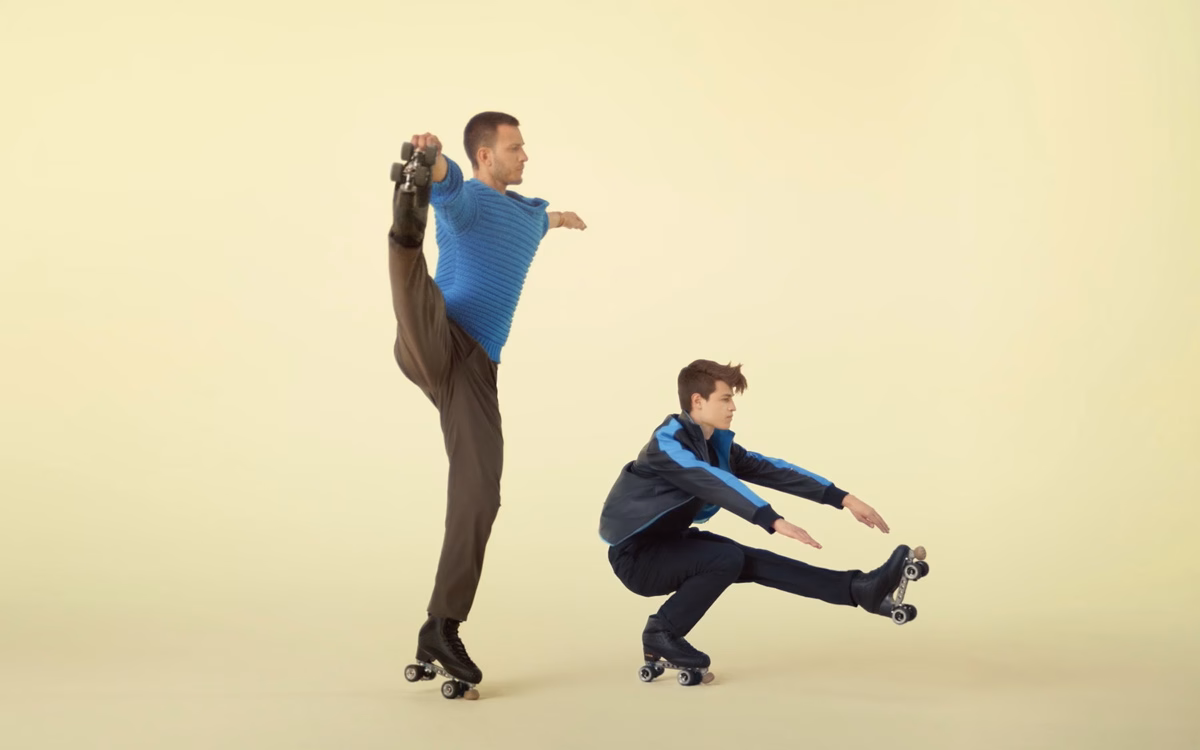 Two roller skaters pose on a soft yellow background, one balancing on one leg while stretching, the other crouched.