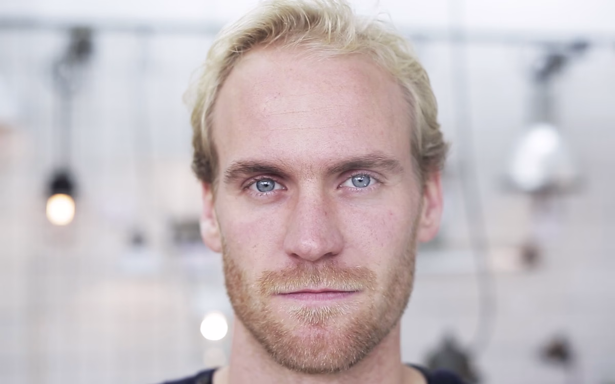 A man with light blonde hair and blue eyes gazes directly at the camera against a softly blurred background.