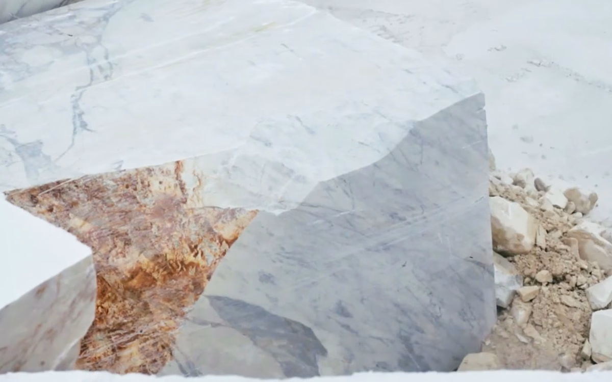 A close-up of a large marble block, showcasing white and warm brown veining amidst rough surrounding stone.