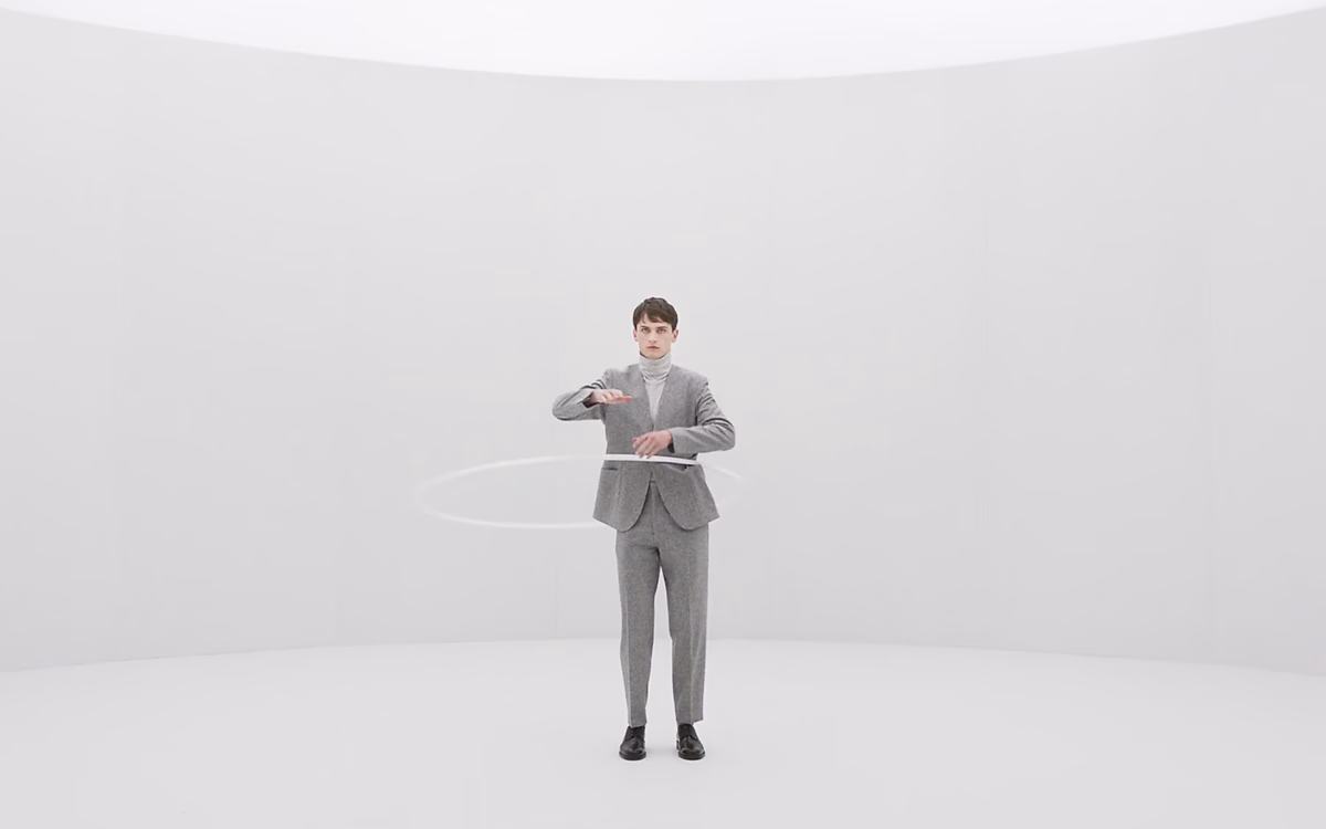 A man in a grey suit performs with a hula hoop in a minimalist, white-walled space.