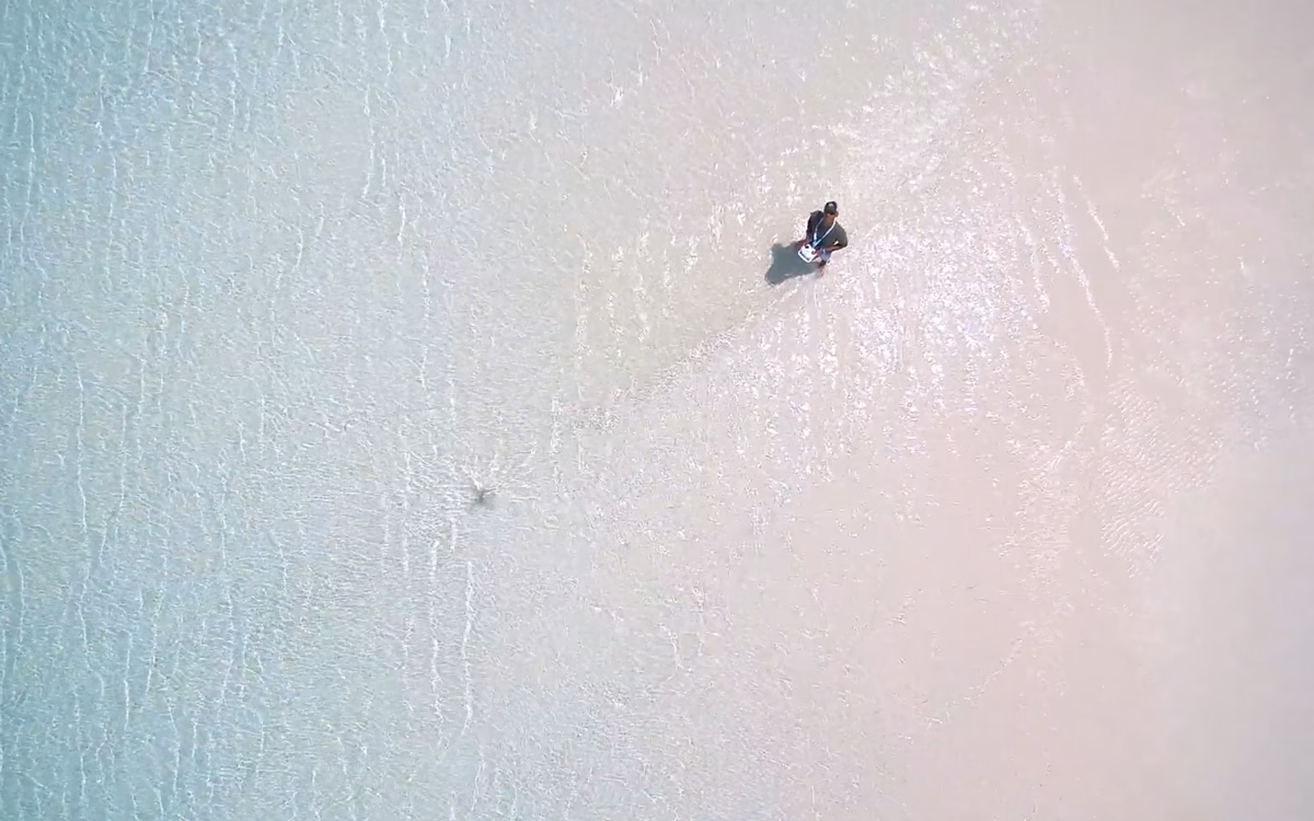 Aerial view of a person standing in shallow, clear turquoise water on a sandy beach, surrounded by gentle waves.
