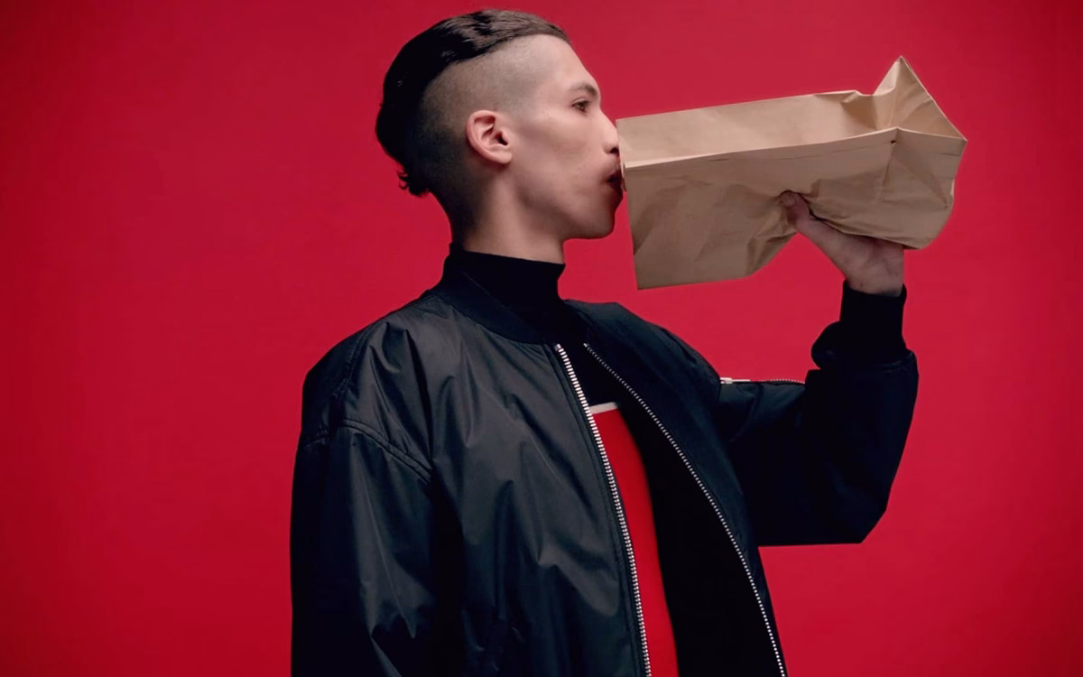 A person in a black bomber jacket holds a brown paper bag against a vibrant red background.