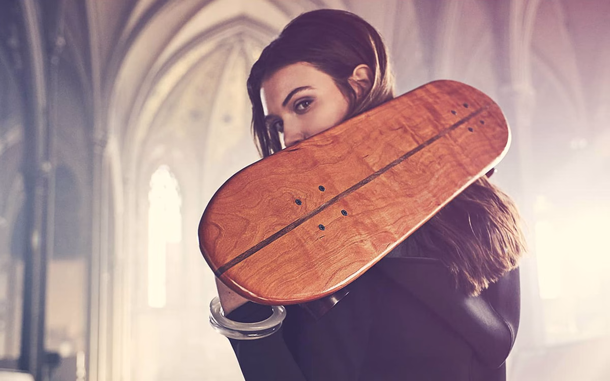 A woman with long hair holds a wooden skateboard against her shoulder, set in a softly lit, gothic interior.