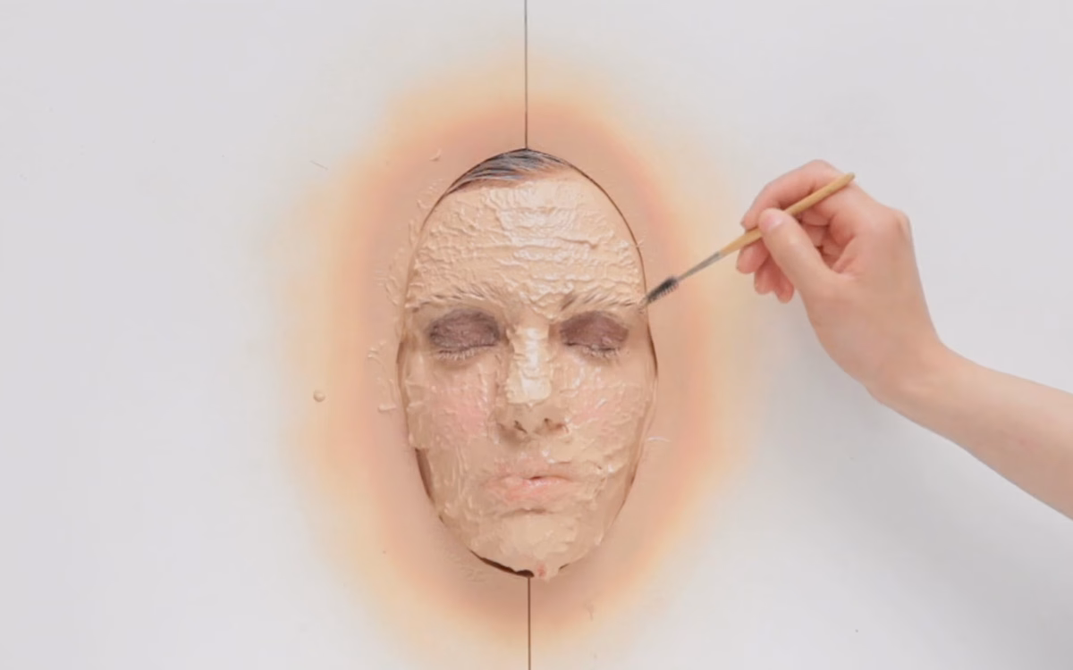 A close-up of a semi-realistic face sculpture being painted with beige material, showcasing textured features.