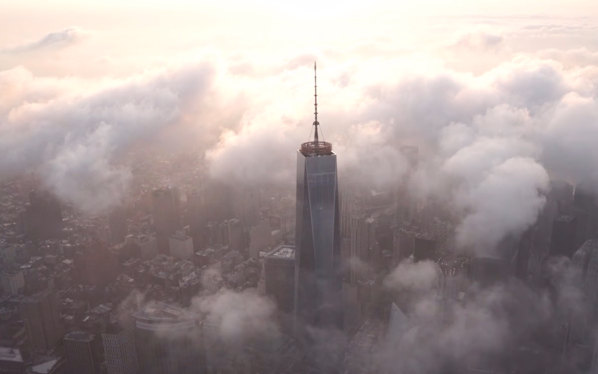One World Trade Center rises above a cityscape shrouded in soft clouds during a warm, hazy sunrise.