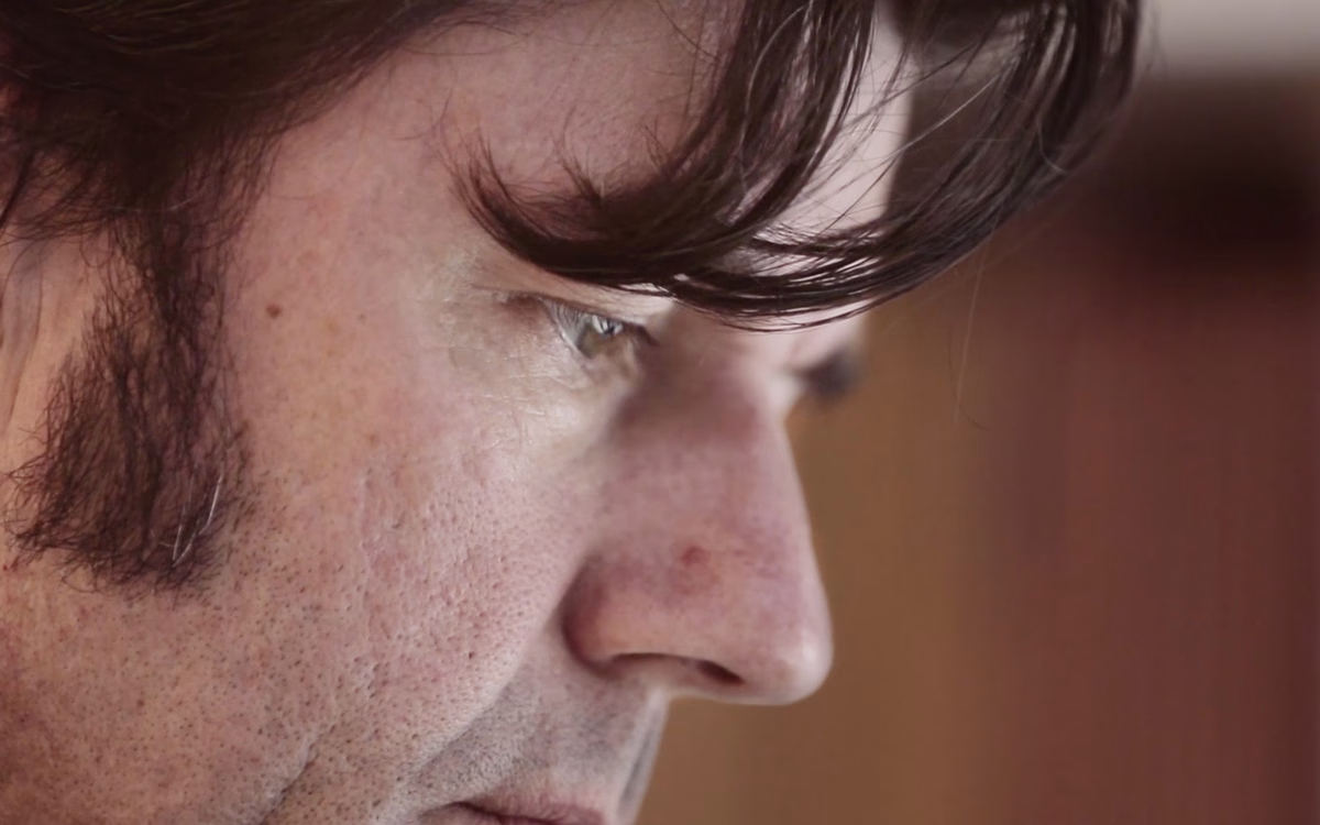 A close-up profile of a man with dark hair and a focused expression against a blurred background.