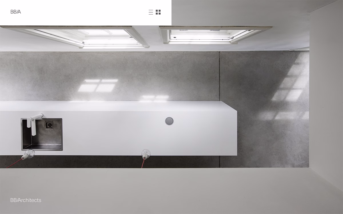A minimalist kitchen space features a long white countertop and stainless steel sink, with natural light casting shadows.