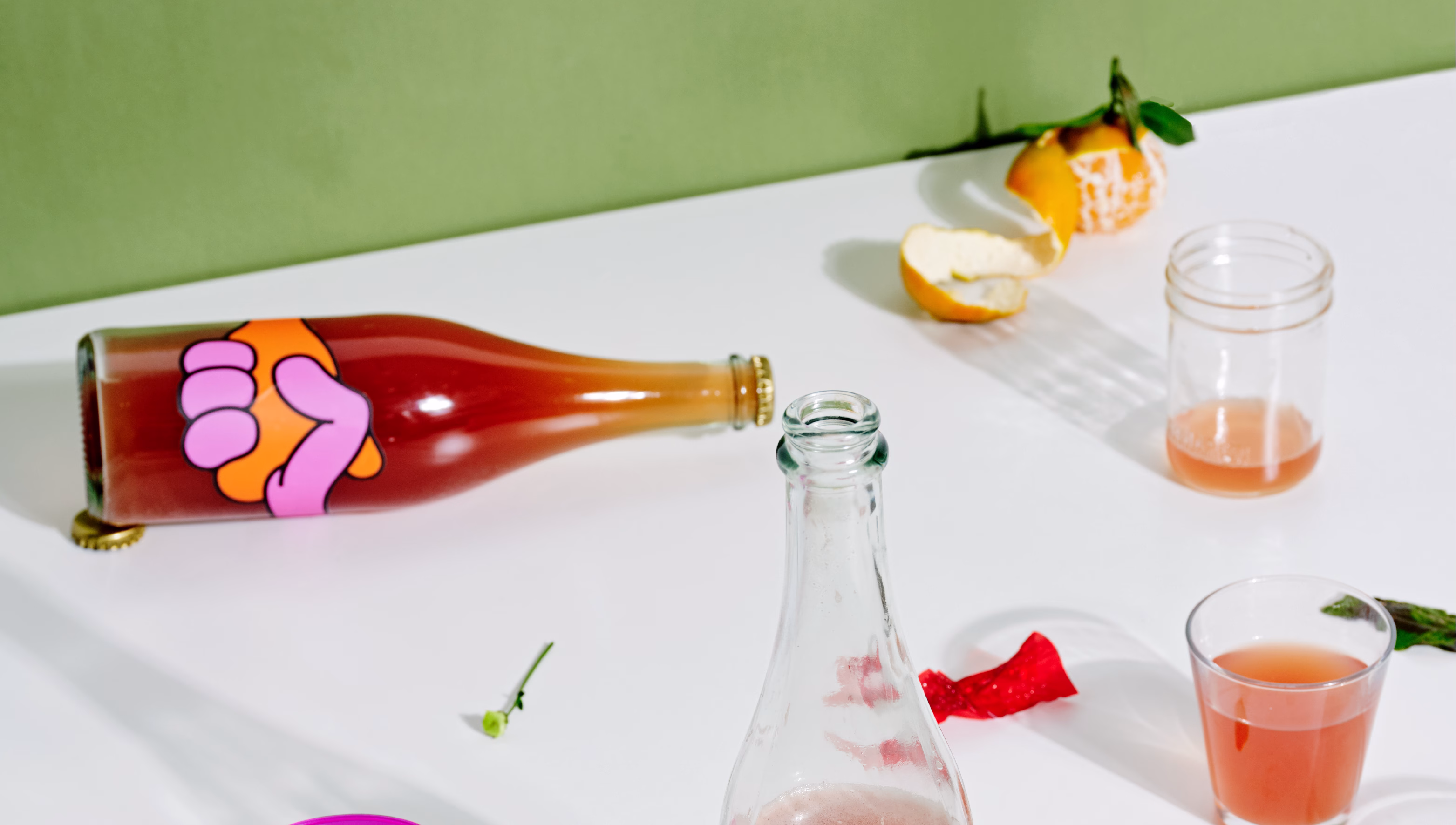 A tipped bottle with a colorful label sits beside a glass jar and citrus peels on a white table against a green backdrop.