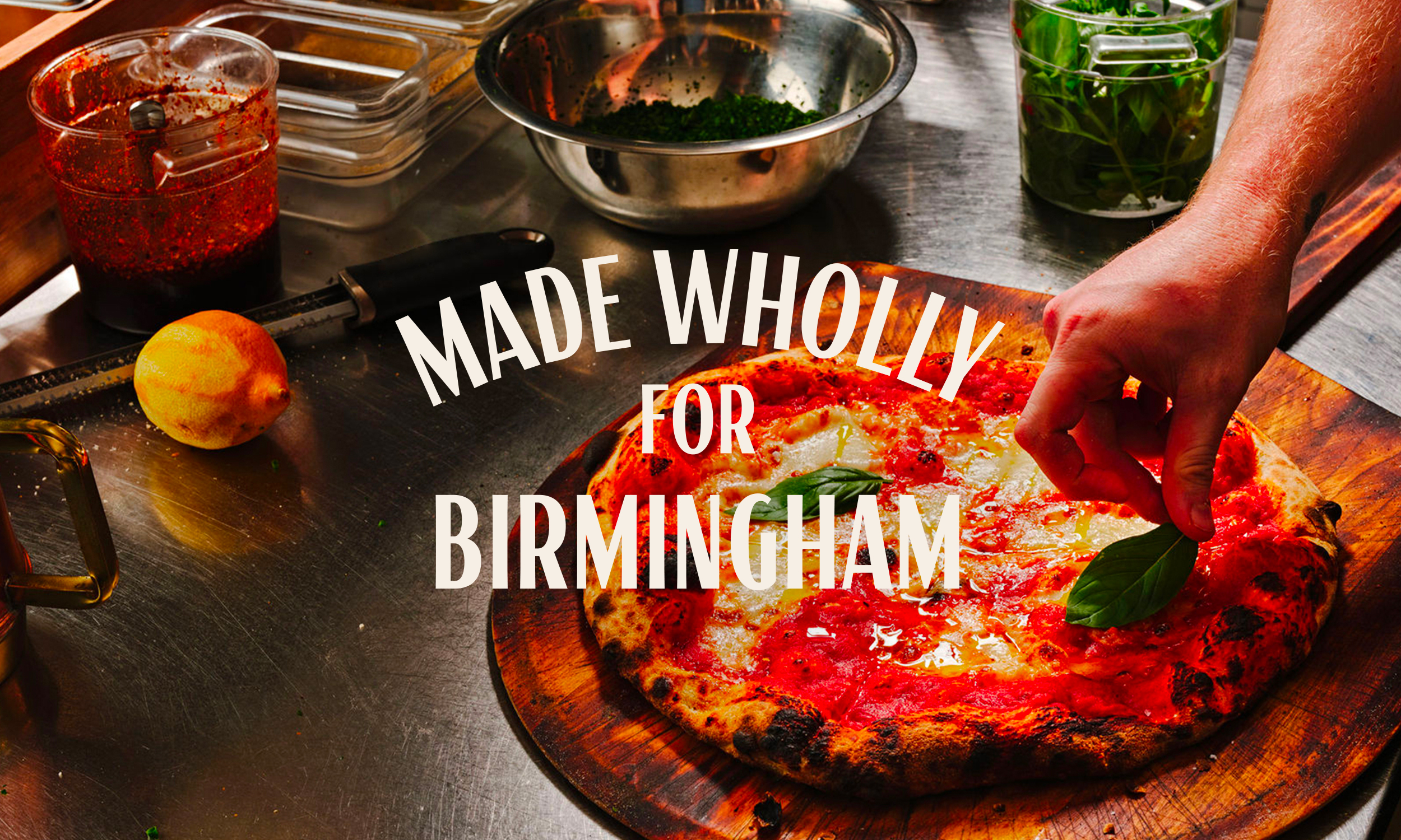 A hand garnishing pizza with basil; text reads "Made wholly for Birmingham."