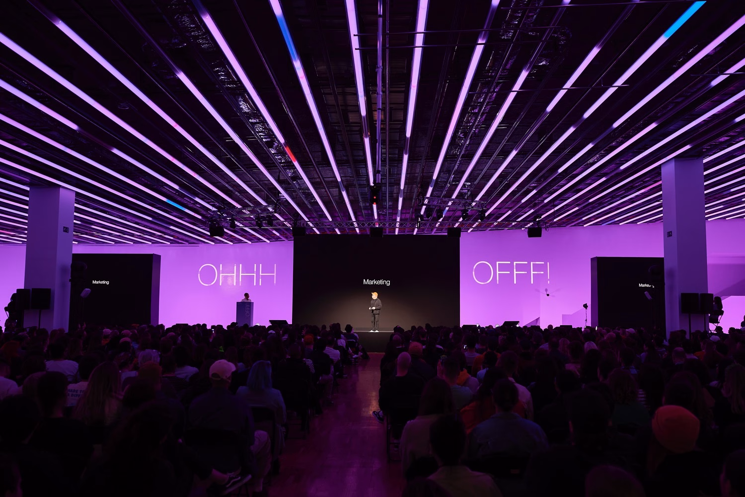 A speaker stands on stage in front of a large audience, illuminated by vibrant purple lighting with "Marketing" displayed behind.