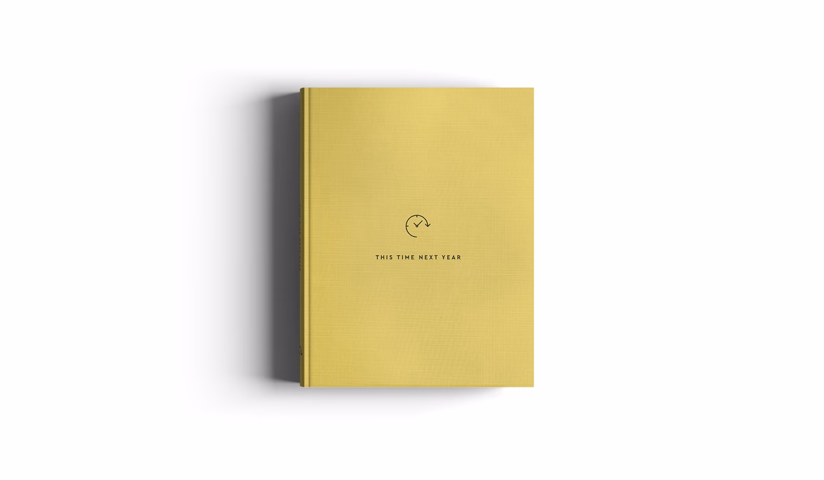 A yellow hardcover book lies flat, featuring the text "THIS TIME NEXT YEAR" and a simple icon on the cover.
