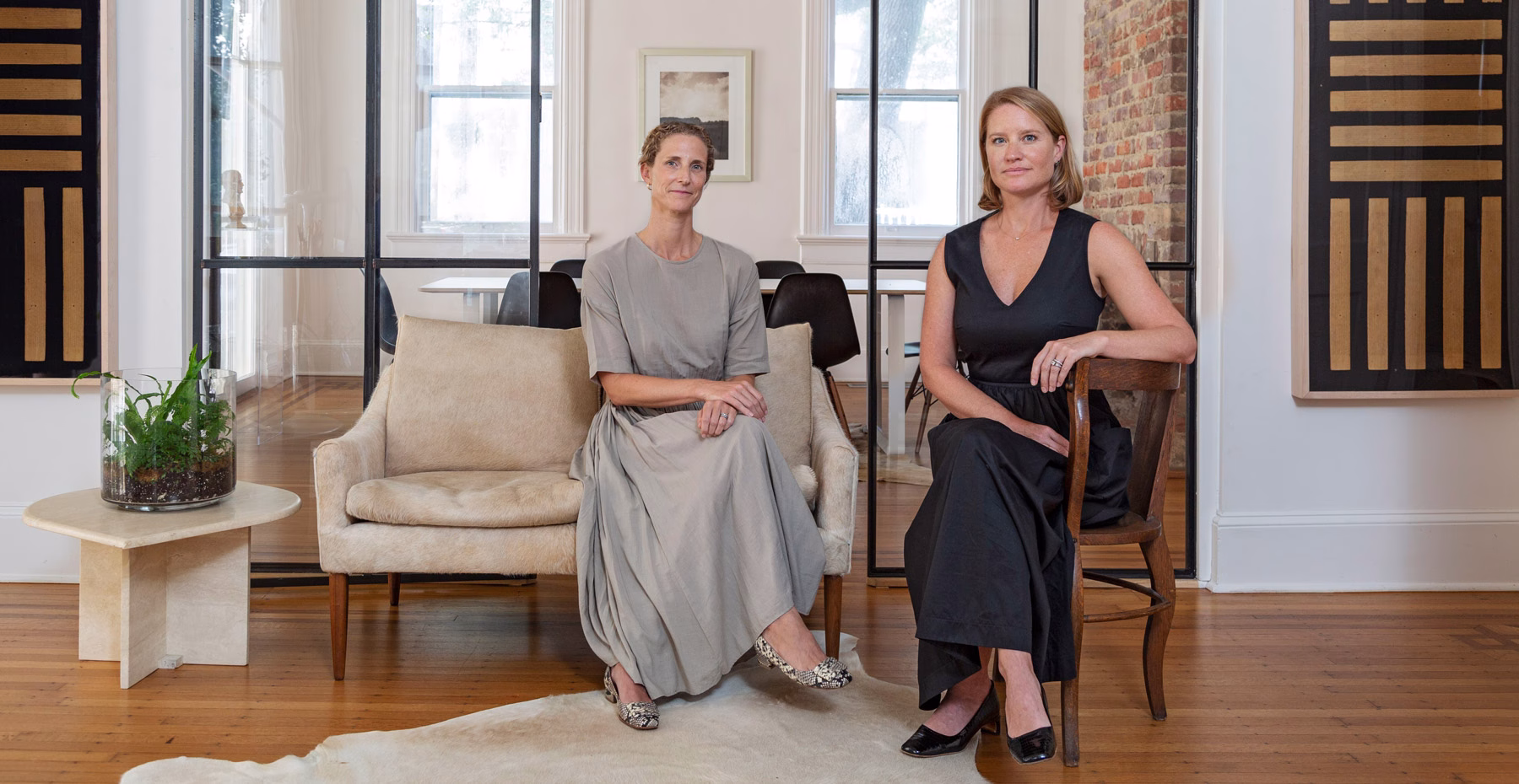 Two women sit on a beige sofa in a modern interior, framed by glass walls and minimal artwork. Natural light fills the room.