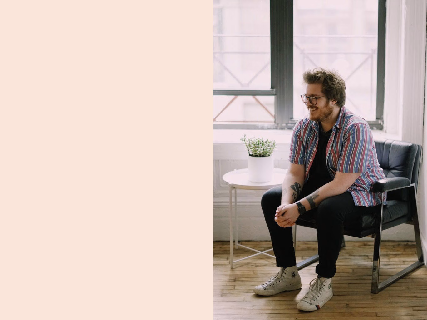 A person with glasses sits casually in a black chair, wearing a striped shirt and white sneakers, smiling near a plant.