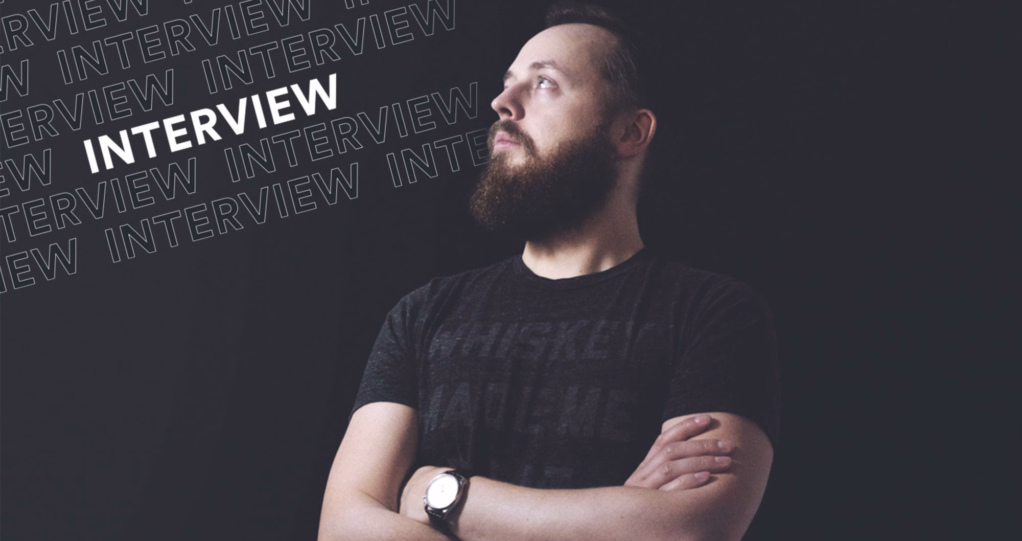 A man with a beard poses thoughtfully against a dark background, wearing a black shirt with a watch on his wrist.