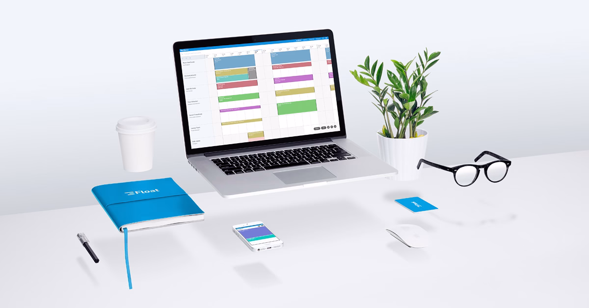 A laptop displays a colorful calendar, accompanied by a blue notebook, coffee cup, glasses, and a plant on a white desk.