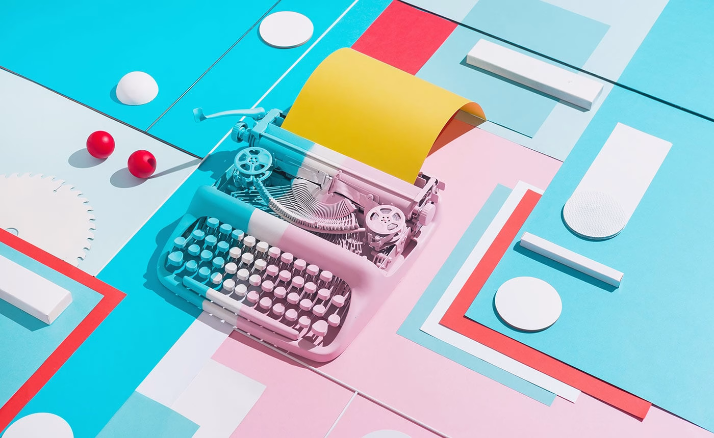 A pastel pink typewriter sits on colorful geometric paper, featuring yellow paper emerging from the top and red balls nearby.