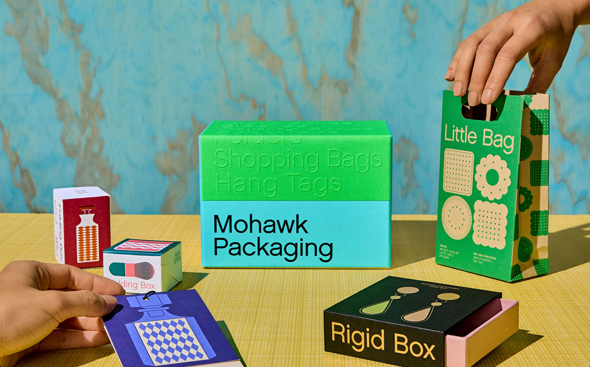 Colorful packaging boxes and bags with hands arranging them on a yellow surface.