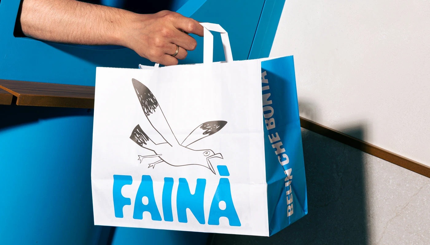 Hand holding a paper bag with a bird illustration and the text "FAINÀ."
