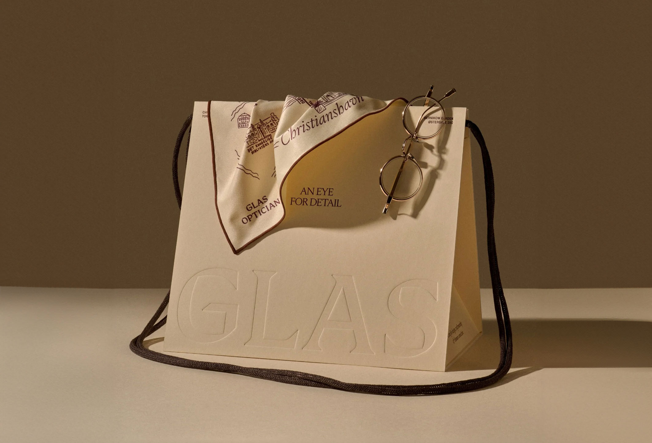 A paper bag with "GLAS" embossed, a cloth, and round eyeglasses on top.