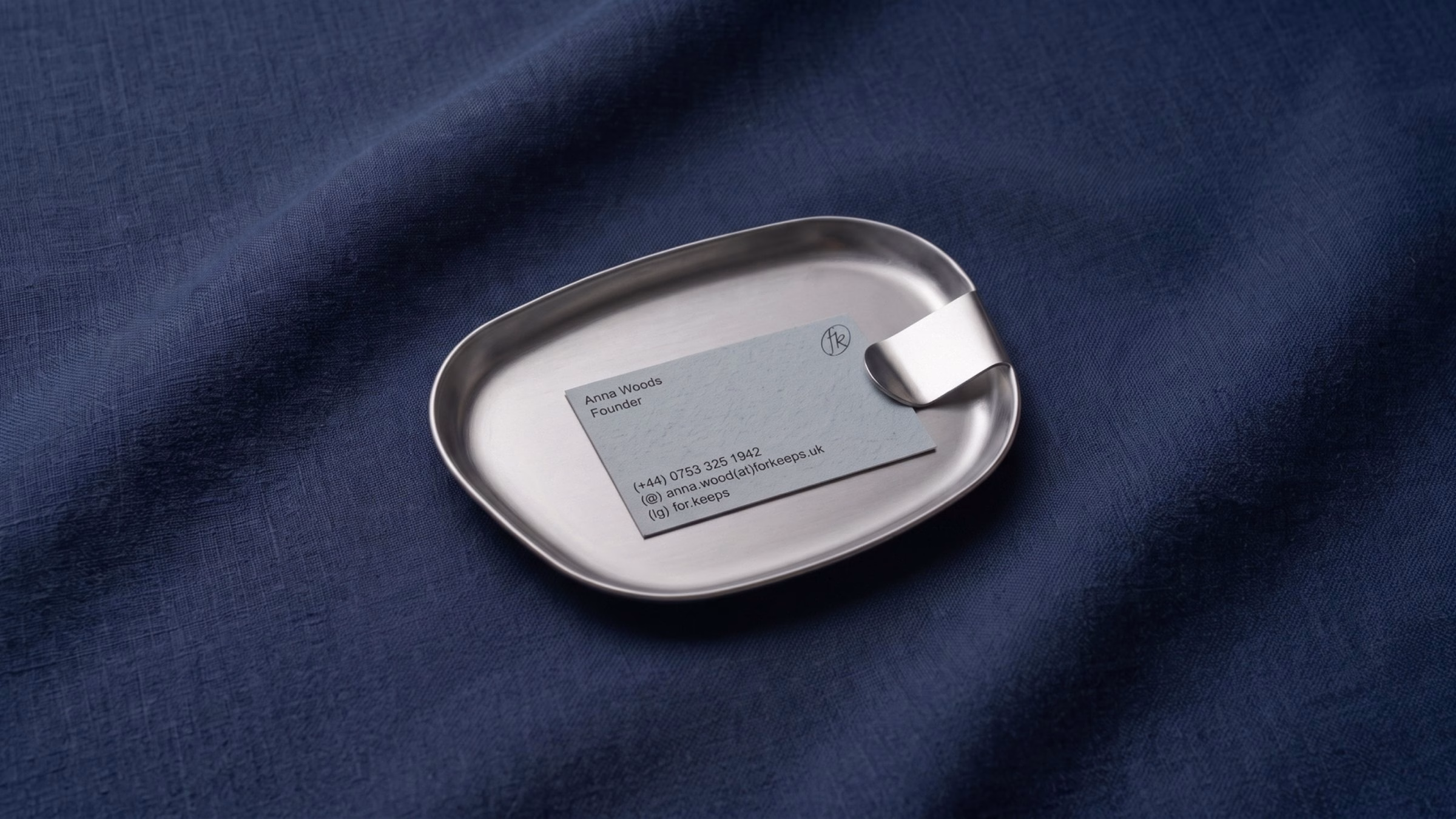 A pale blue business card rests on a silver tray atop a deep blue textured fabric, reflecting For Keeps’ brand identity.
