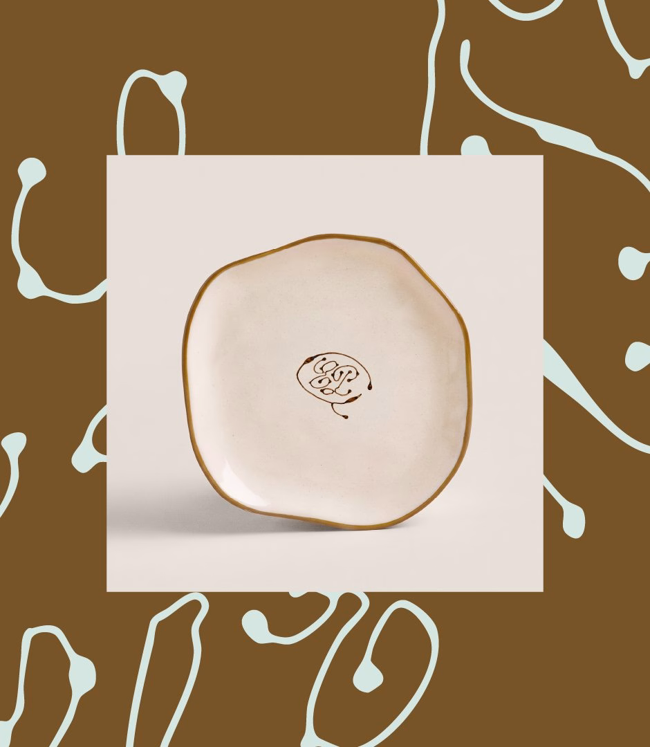 A handmade plate with an organic shape and a brown logo, set against a contrasting brown and light blue patterned background.