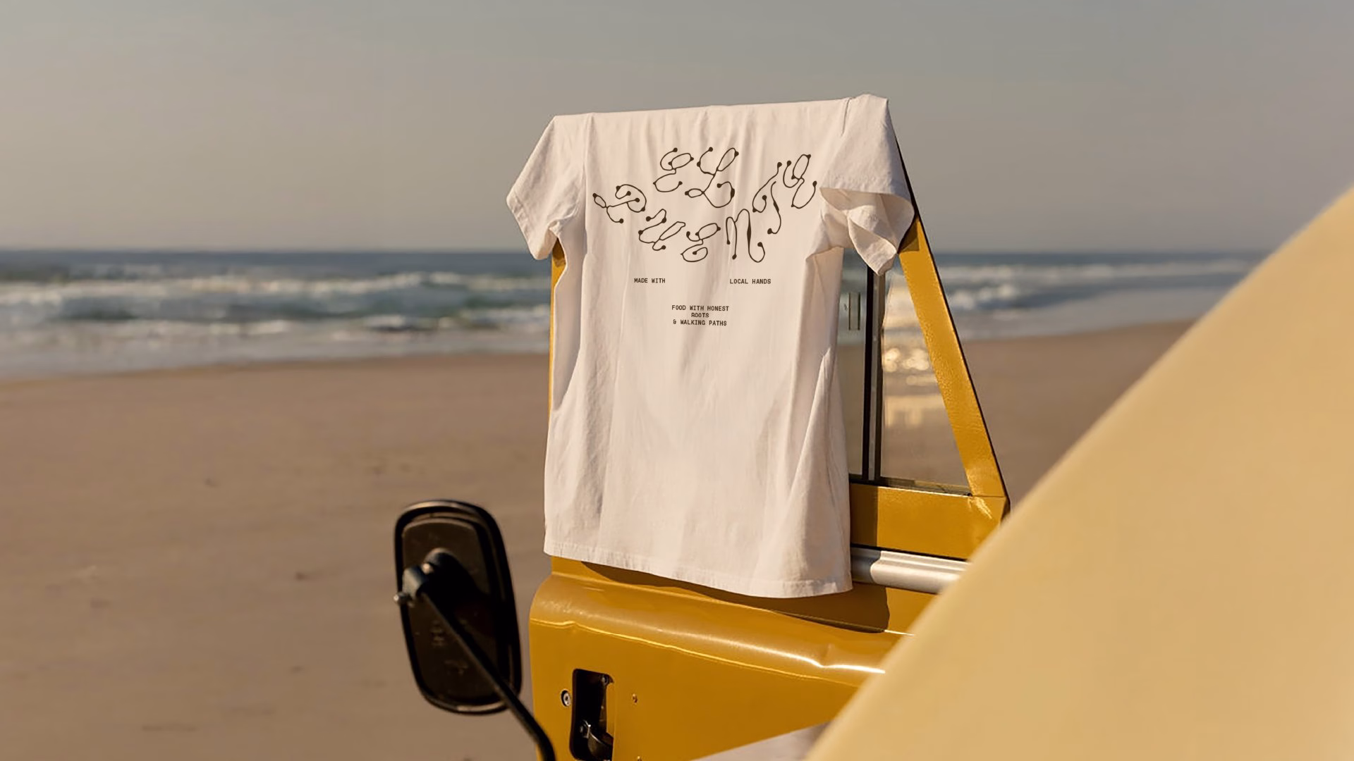 A white t-shirt with wavy typography hangs from a yellow vehicle, set against a sandy beach and ocean backdrop.