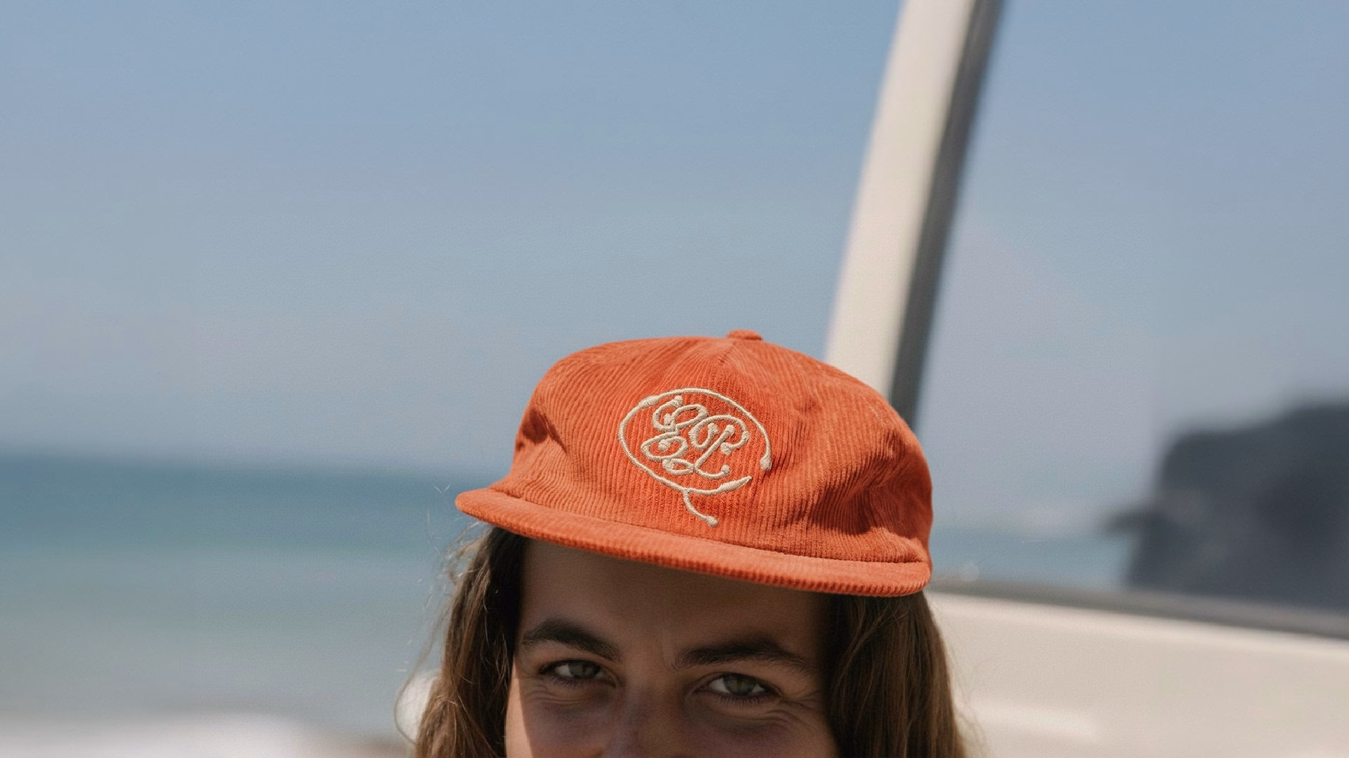 An individual wears an orange corduroy cap featuring a white logo, set against a beach backdrop.