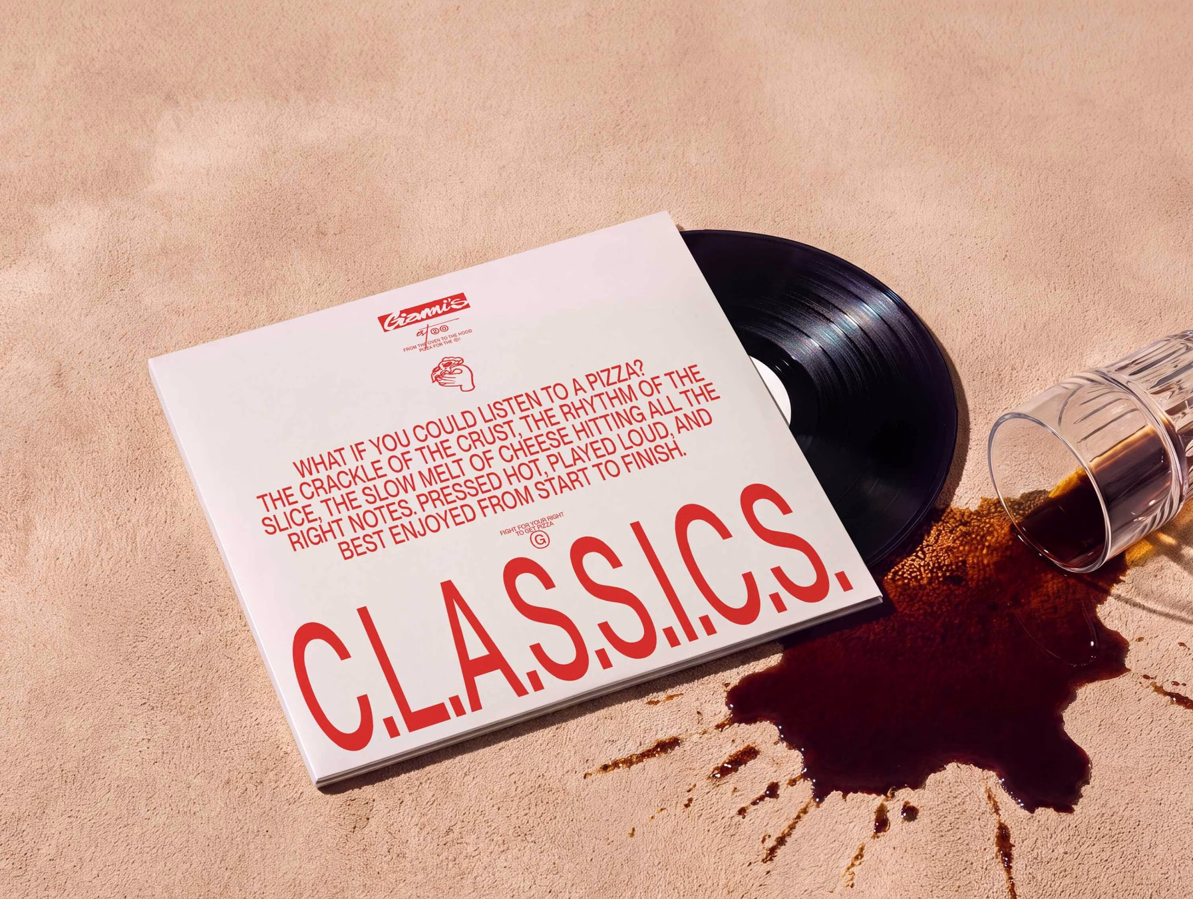 A vinyl record next to a white album cover with bold red typography, resting on a textured beige surface with spilled coffee.