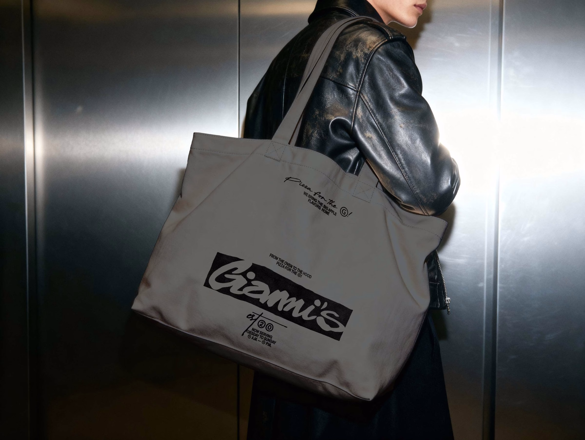 A model in a black leather jacket carries a beige tote bag featuring the Giannis logo and text in bold black typography.