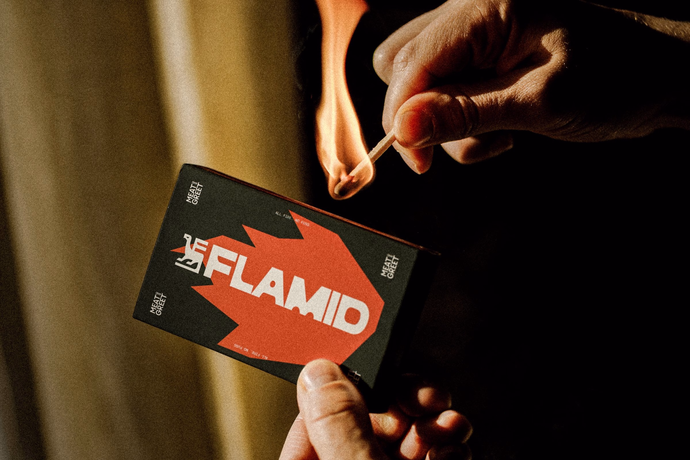 A hand ignites a match near a black and red box bearing the Flamid logo, set against a blurred background.