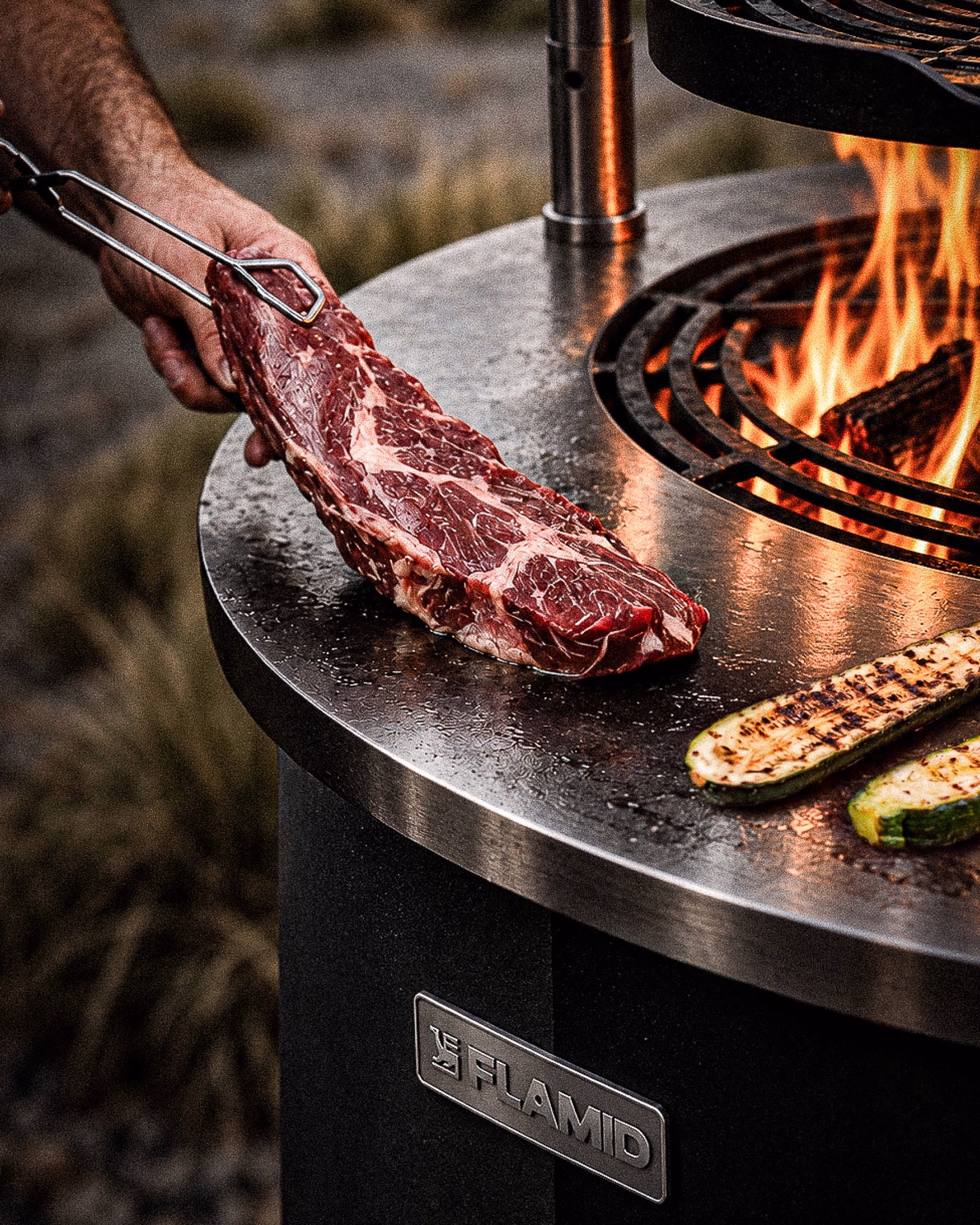 A hand holds a raw steak above a steel grill branded with the Flamid logo, featuring flames and grilled vegetables nearby.