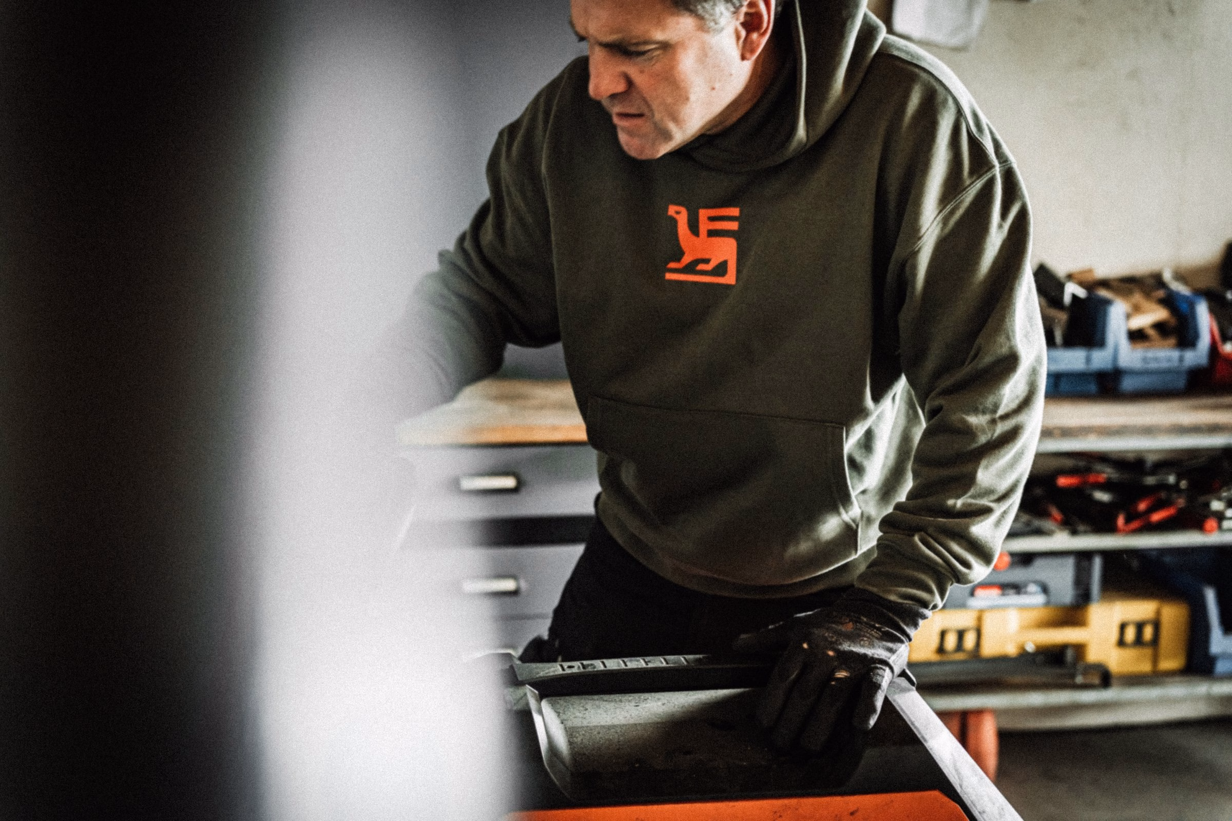 A man wearing a dark green hoodie with an orange bird-in-the-F logo works in a workshop, focused on a black tool.