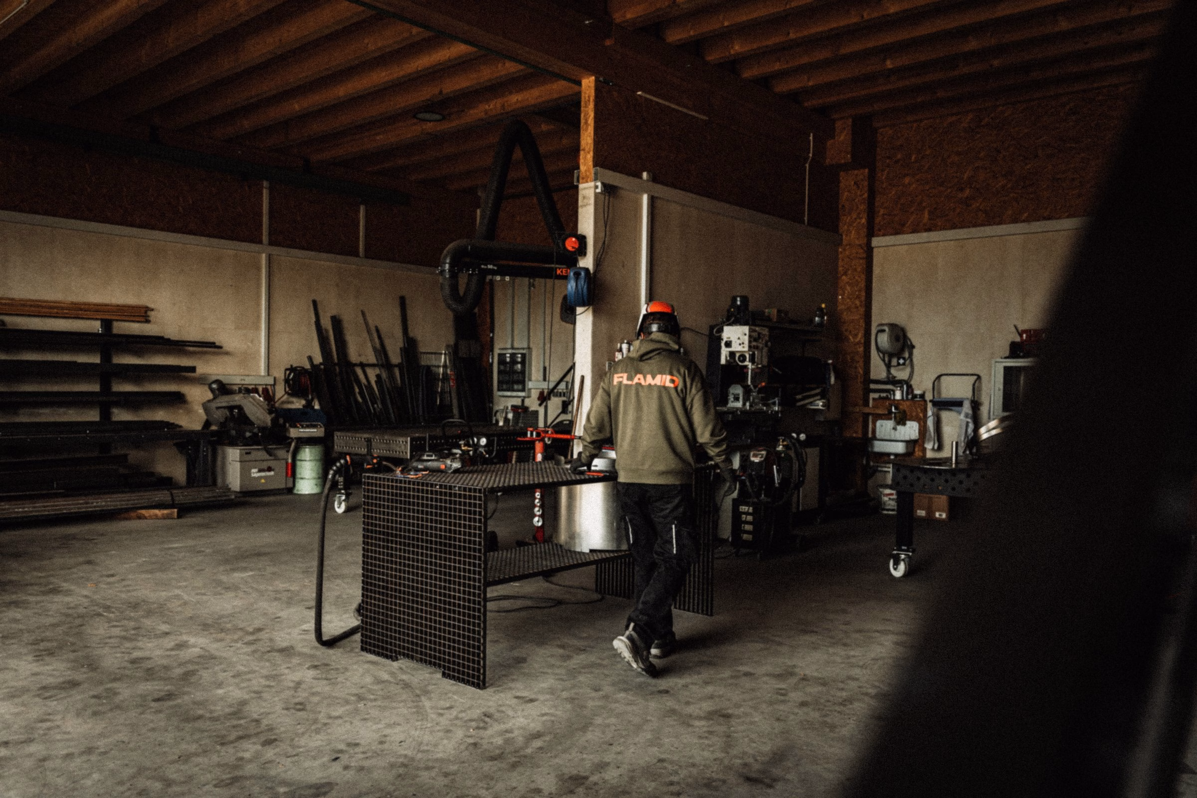 A person in a green FLAMID hoodie walks past a black grid worktable in a dimly lit workshop filled with tools.