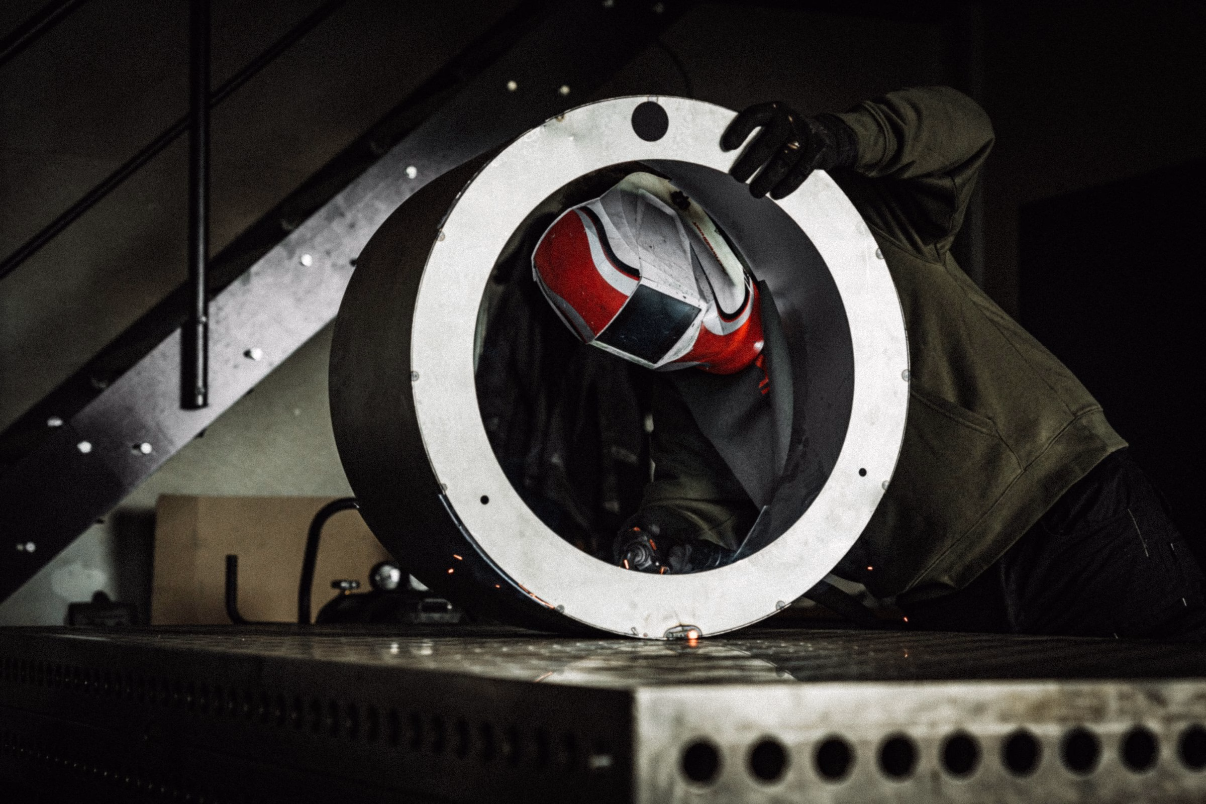 A welder works on a circular steel grill component, focusing intently through a safety helmet with a red visor.