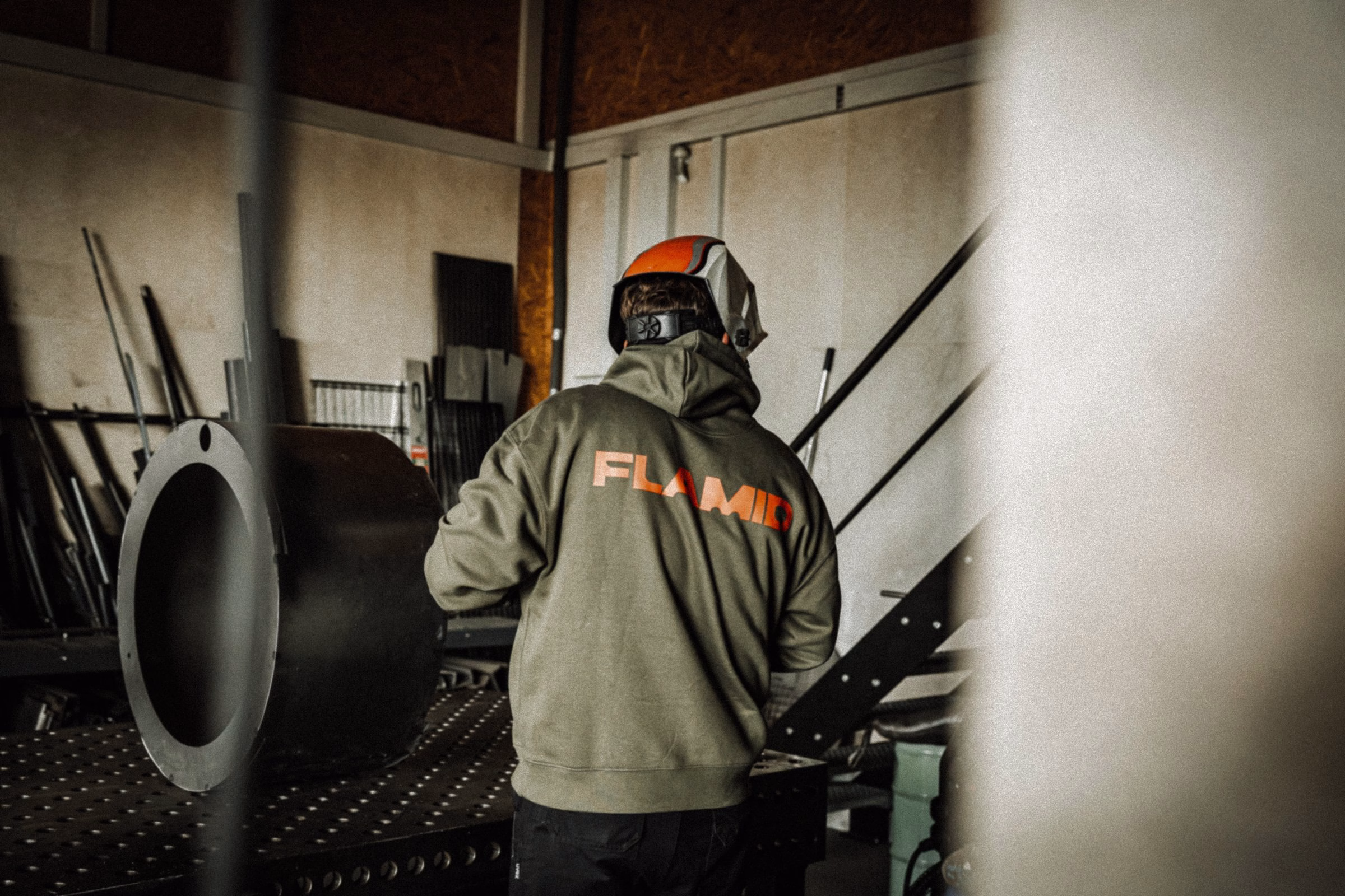 A person in a green hoodie with "FLAMID" printed on the back stands near a large steel round object in a workshop.