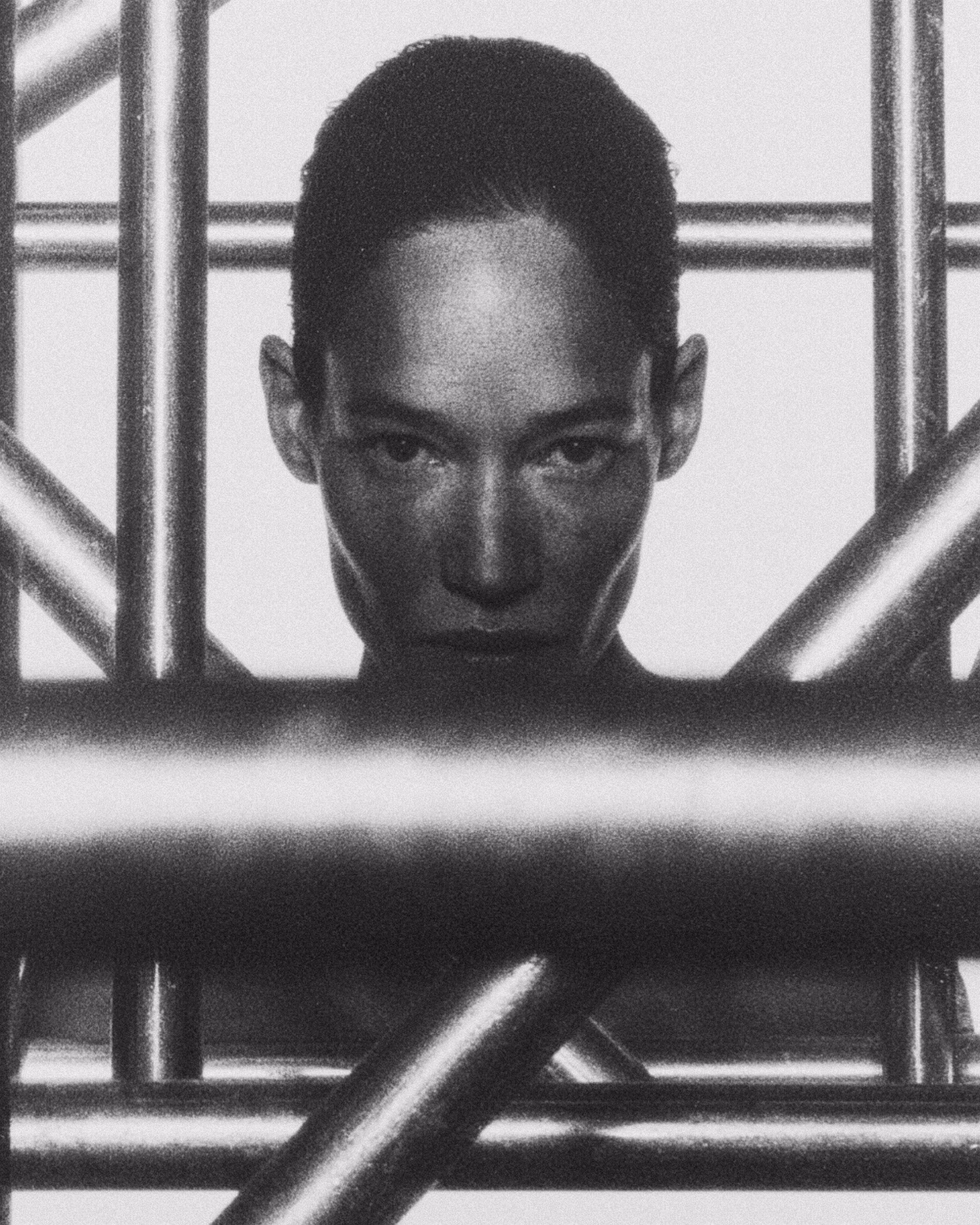 A close-up of a person's face framed by metallic grid-like structures, evoking tension in monochrome tones.