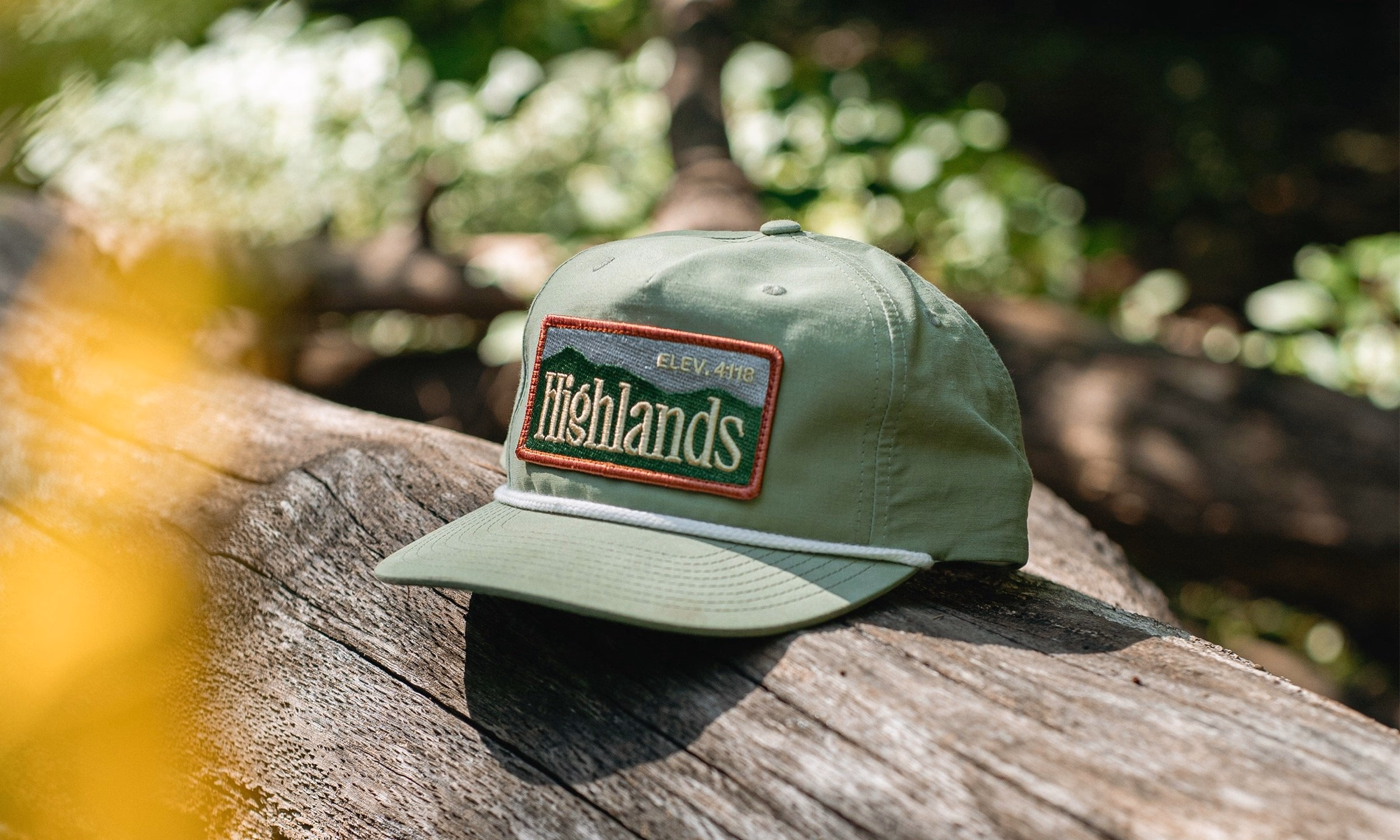 A green cap with a patch featuring the word "Highlands" rests on a weathered log, surrounded by greenery.