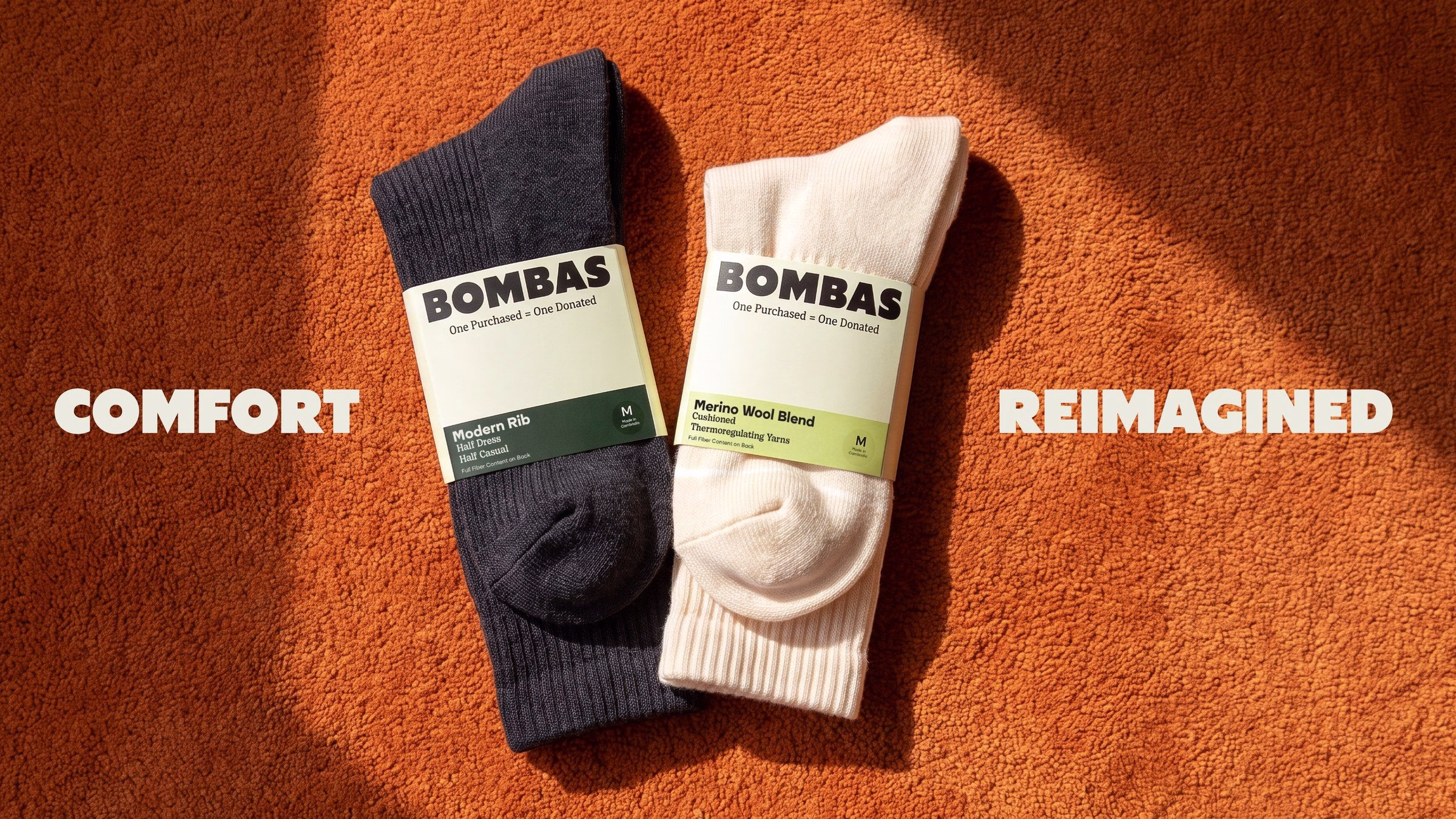 Two pairs of Bombas socks, one dark navy and one cream, displayed on an orange textured surface, showcasing their packaging.