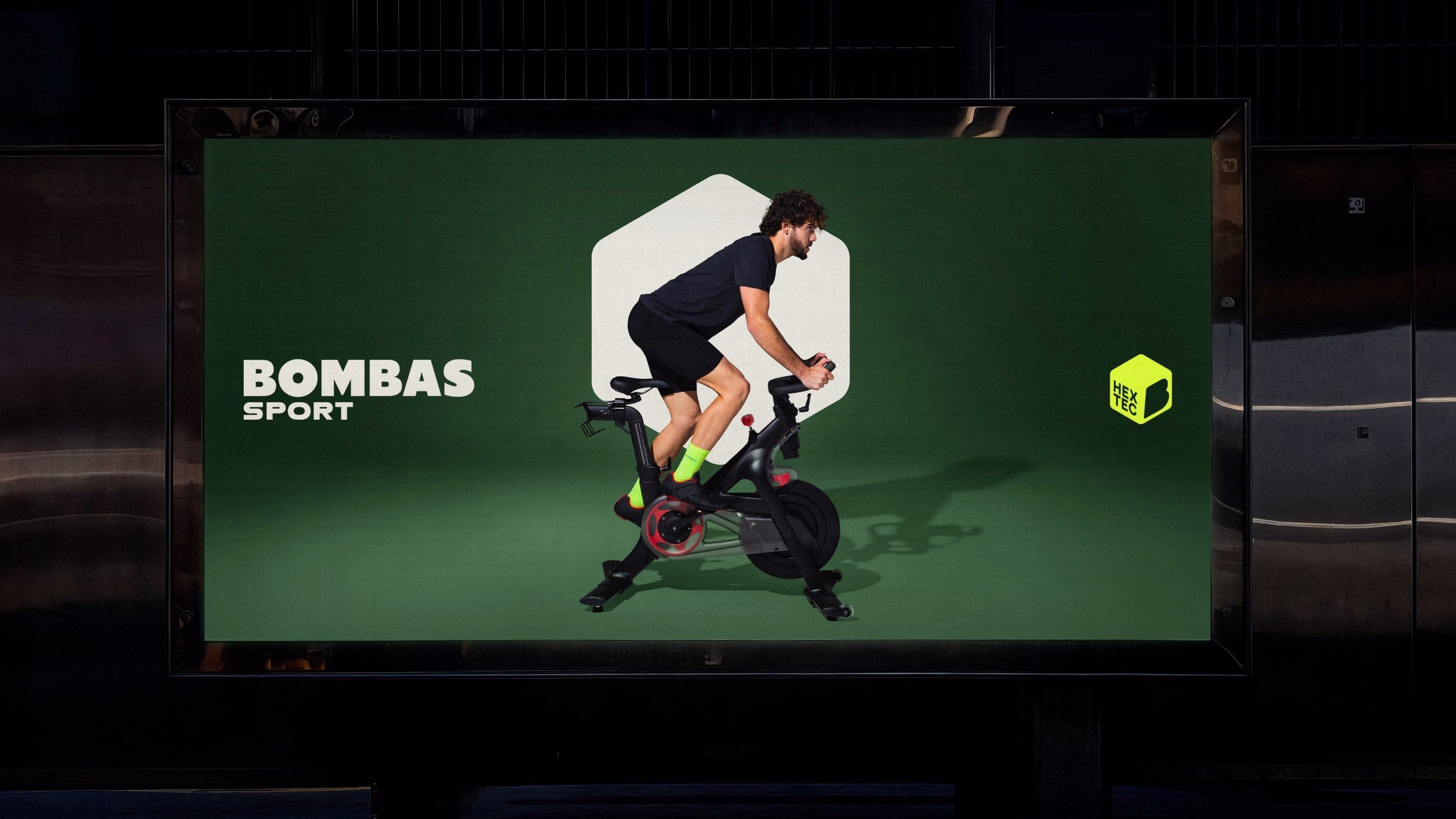 A cyclist in activewear pedals on a stationary bike against a green background with bold "Bombas Sport" typography.