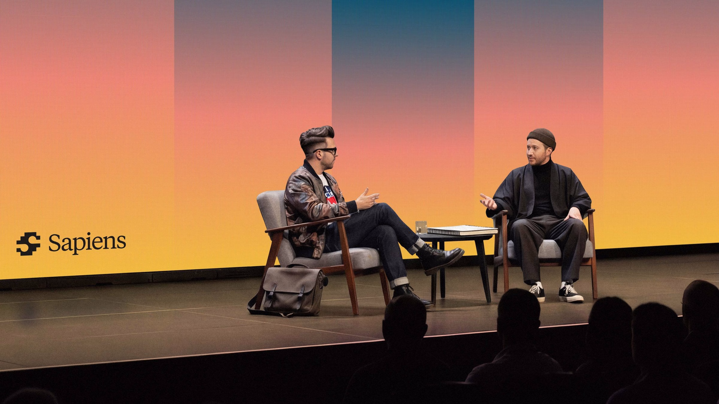Two speakers engaged in discussion on stage with a colorful gradient backdrop, representing Sapiens’ modern storytelling.